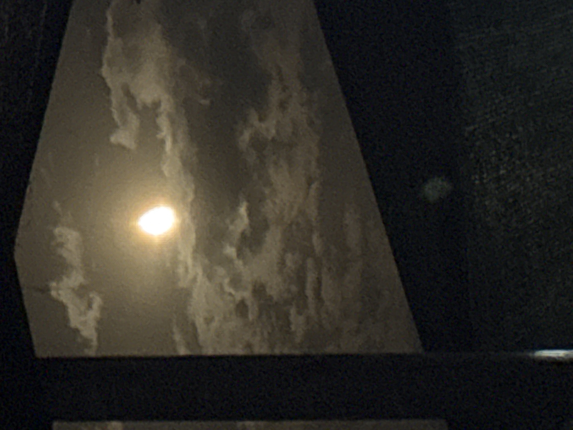 The image depicts a nighttime scene with a bright, luminous moon dominating the sky. The moon is partially obscured by scattered clouds, creating a dynamic interplay of light and shadow across the celestial expanse. The lower portion of the image is darker and less defined, suggesting the presence of a foreground element, possibly a window frame or a dark surface, that partially obstructs the view of the sky. The overall mood is serene and atmospheric, with the moonlight casting a soft glow on the clouds. The location of Vostok, Kyrgyzstan, is not discernible from the visual information provided in the image.