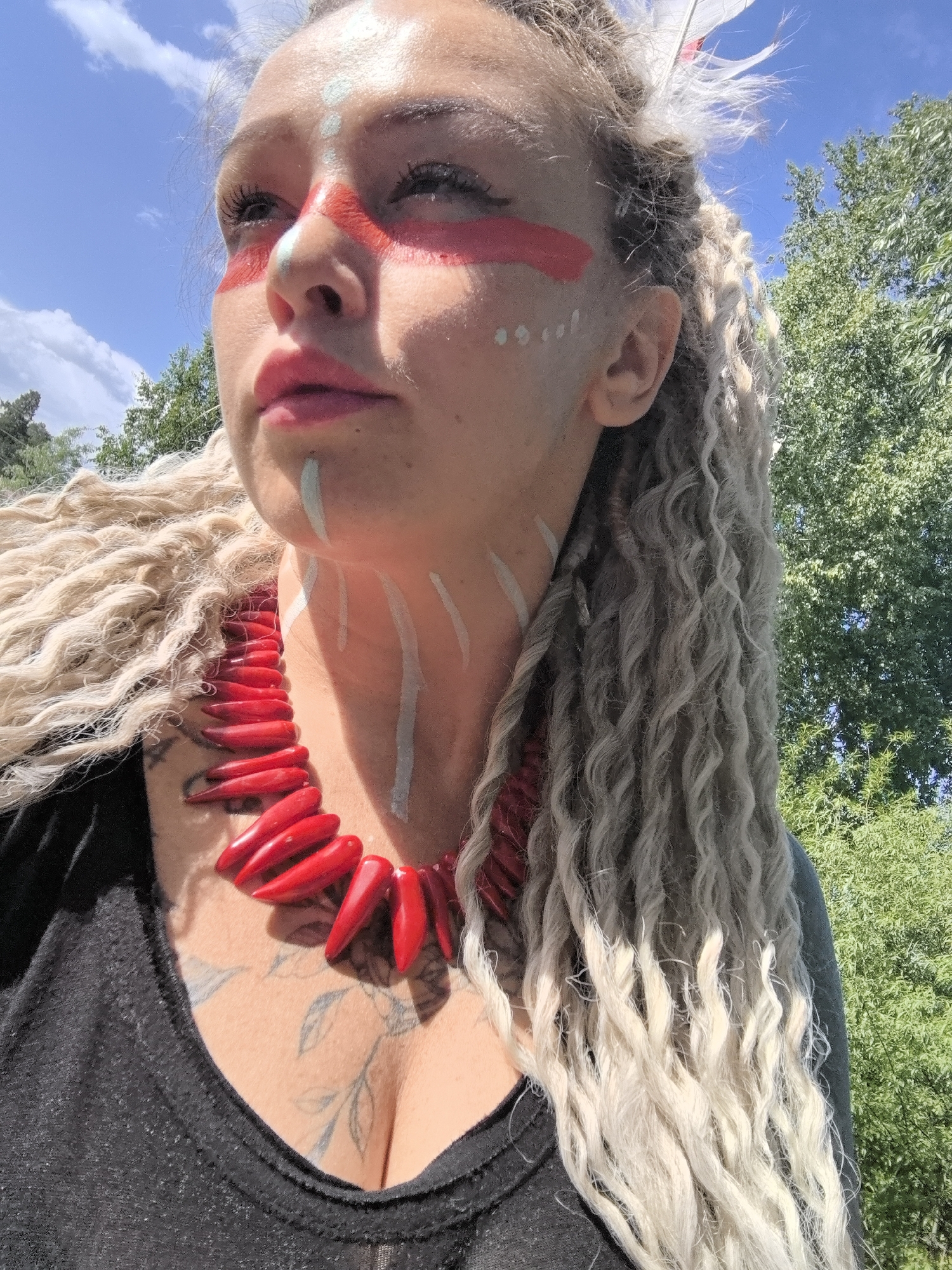 A medium shot captures a person wearing face paint reminiscent of indigenous tribal styles. Red paint adorns the eye area, and a series of white dots mark the temple. A vertical white line extends from the lower lip. The neck is painted with vertical white lines. The person wears a large red necklace of shaped beads. Blonde dreadlocks frame the face and extend down the sides of the torso. A white feather is visible in the hair. The person wears a black shirt. A leafy branch tattoo is visible between the breasts.
The background features a partly cloudy sky, greenery, and trees. There are no identifiable locations or specific places in the image.