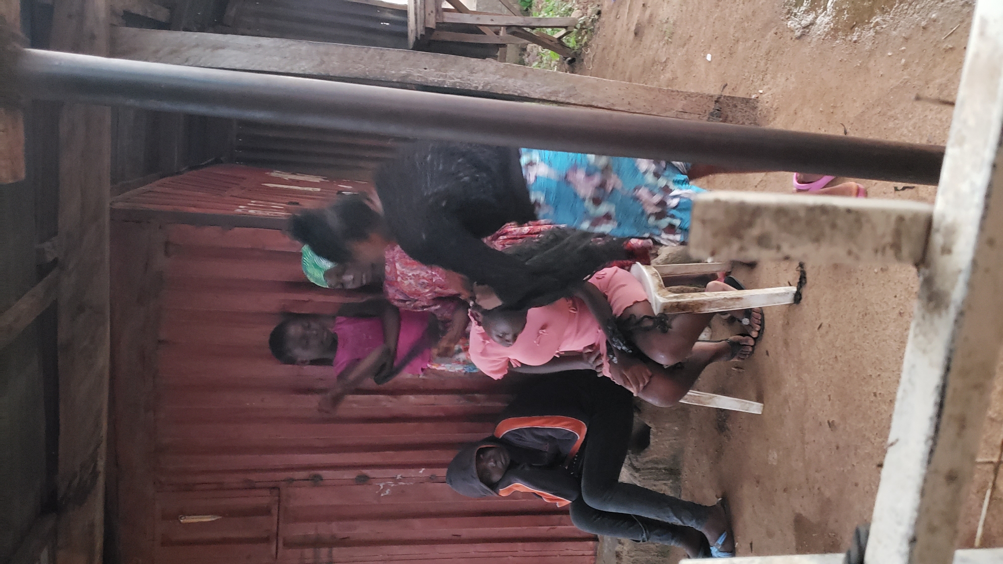 A group of young women and girls are gathered in an outdoor, makeshift shelter in Narayi, Nigeria. The structure appears to be made of corrugated metal walls and a wooden roof. In the foreground, a young woman with dark, long hair is seated on a white plastic stool. Another young woman, wearing a pink dress, is seated next to her, also on the stool, with her head bowed. A third young woman, wearing a black hoodie and dark pants, is seated on a raised concrete step to the left, leaning her head on her hand. Two other young women are standing behind the seated individuals. One wears a pink sleeveless top and a patterned skirt, and the other wears a patterned dress and a green headscarf. The ground is dirt and sand, and there is some scattered debris. The overall scene suggests a casual gathering or perhaps a hair styling session within a modest urban environment.
