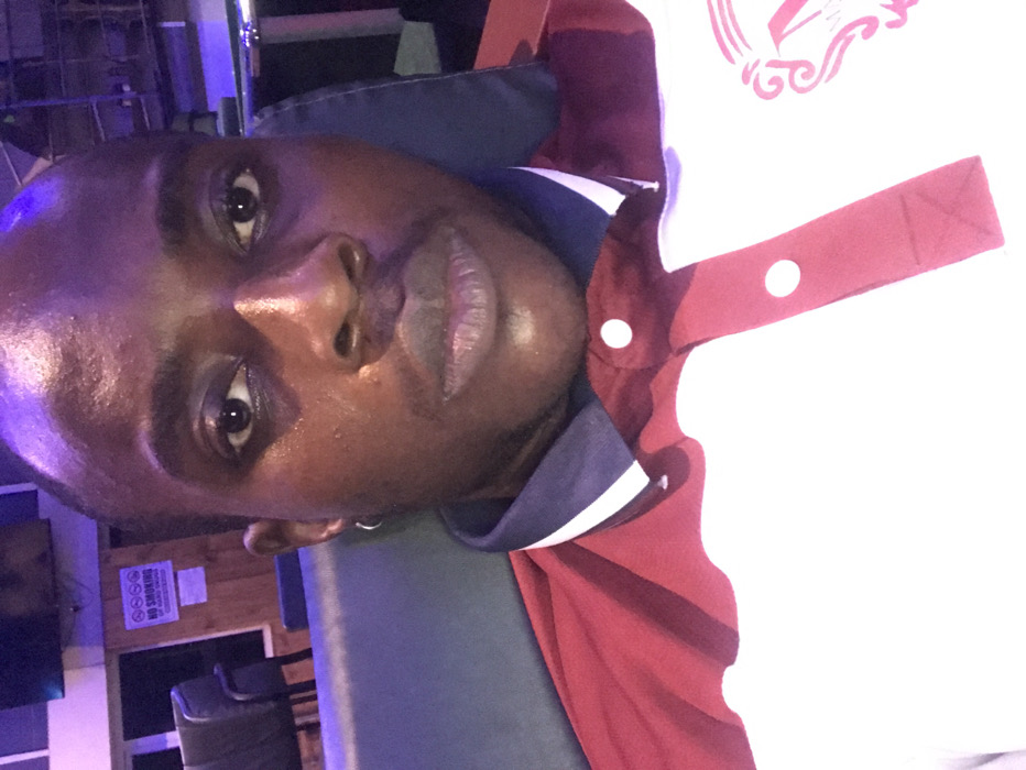 A male individual with dark skin, short dark hair, and a neutral expression is captured in a close-up, front-facing image, looking directly into the camera. He is wearing a short-sleeved polo shirt with a white main body, a dark blue collar, and dark red panels on the shoulders and button placket. Two white buttons are visible on the placket. A small silver hoop earring is present in his left ear. A partial pink graphic or logo is visible on the lower right side of his shirt.
The setting is an indoor, dimly lit environment, possibly a lounge or similar commercial establishment, characterized by a prominent purple and blue ambient lighting scheme. In the background, dark upholstered seating is visible, along with a segment of a wooden wall. A white sign with red text, featuring a 'NO SMOKING' emblem and additional unreadable text below, is affixed to the wooden wall. A dark rectangular object, appearing to be a monitor or television, is mounted high on the wall in the upper left background. The location is stated as Lekki, Nigeria.