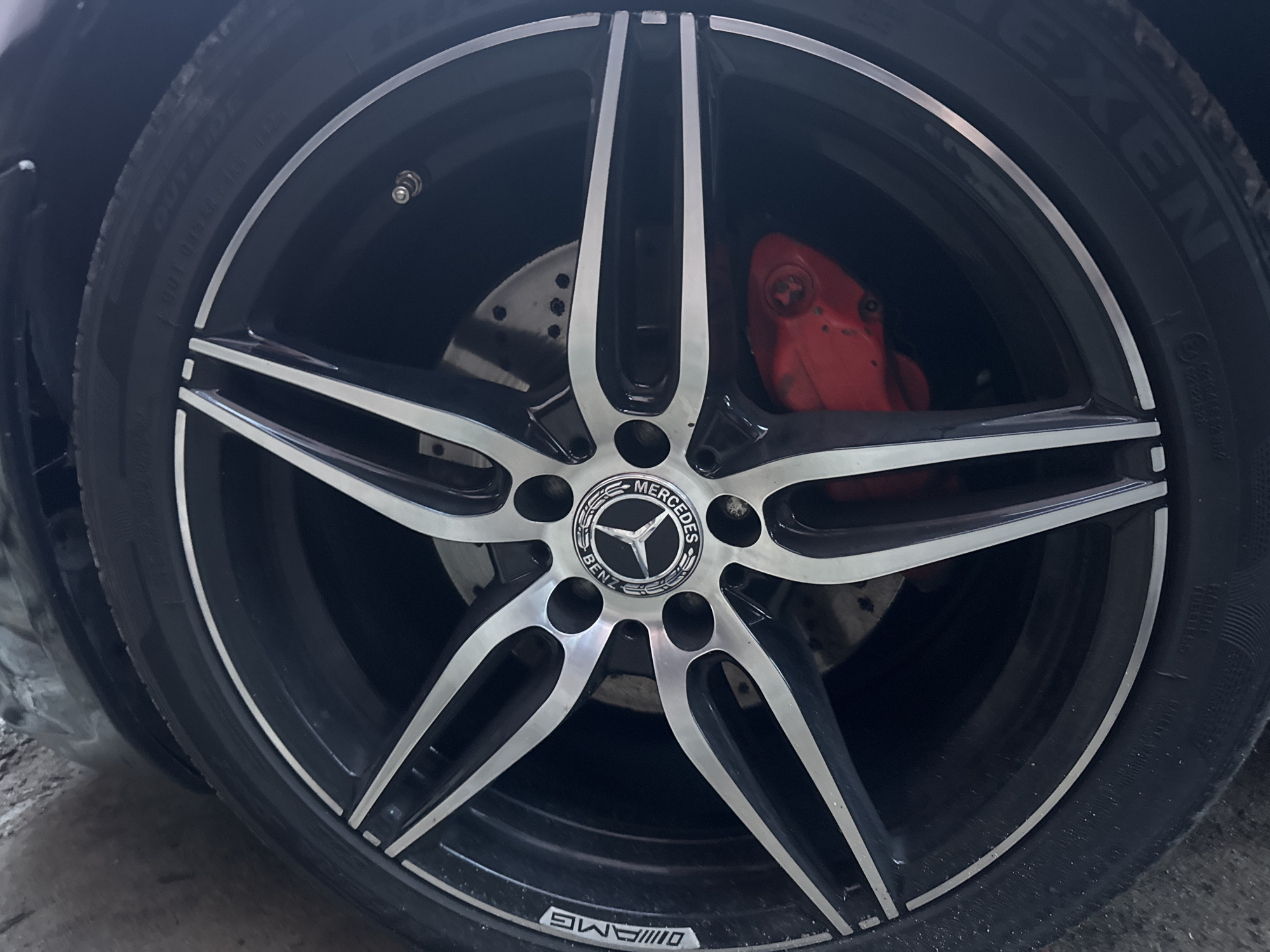 A close-up shot shows a Mercedes-Benz car wheel with a five-spoke silver and black rim. The center cap displays the Mercedes-Benz logo. An AMG logo is visible on the lower part of the wheel. Behind the wheel, a red brake caliper and a drilled brake disc are visible. The tire is black with visible markings and is partially obscured. The photograph appears to be taken indoors, possibly in a garage or workshop. There is no information to confirm the location is in Vratsa, Bulgaria.
