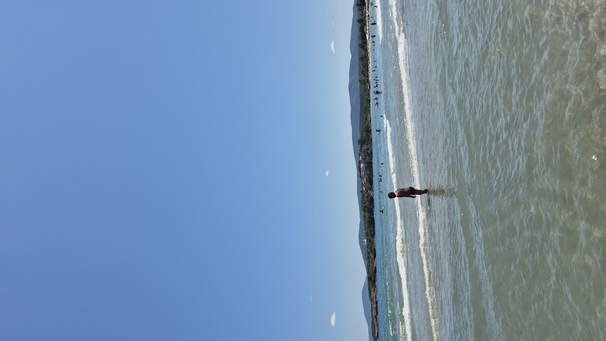 A young boy stands in shallow water at the edge of the Black Sea on a sunny day at the beach in Kiten, Bulgaria. The water is clear and shallow, with small waves gently rolling onto the shore. The boy, wearing black swim trunks, is looking out towards the sea. In the background, other beachgoers are visible swimming and relaxing in the water and on the shore. The coastline features a gentle curve with hills covered in vegetation and buildings in the distance. The sky is a clear, bright blue with a few wispy clouds.