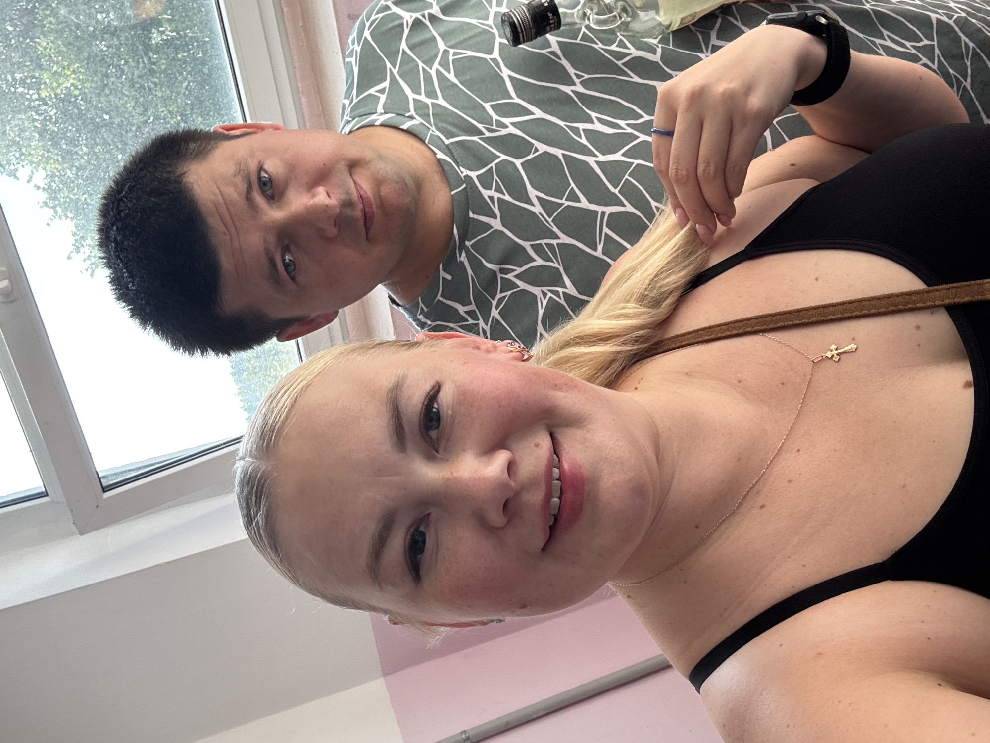 A close-up, selfie-style photograph captures a white woman and a white man in an indoor setting. The woman is in the foreground, to the left, with blonde hair pulled back into a ponytail. She is wearing a black tank top and a gold cross necklace. Her right hand is raised, touching her hair. The man is behind her and to the right, wearing a green and white patterned t-shirt. He is looking directly at the camera with a neutral expression. A clear glass bottle with a black label is visible to the right of the man. The background includes a window with greenery visible outside, suggesting an outdoor scene beyond the window. The interior wall to the left is painted pink and white. The overall lighting is bright and natural. The location is identified as Chaikovsky, Russia.