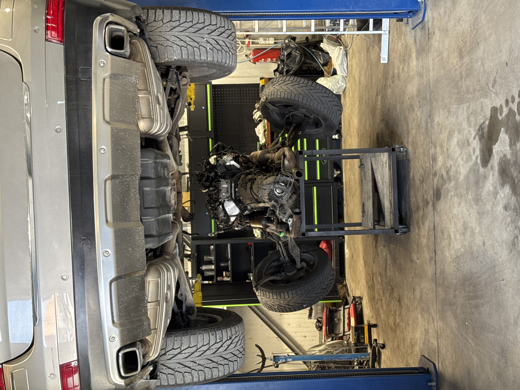 A silver SUV is elevated on a hydraulic lift in a garage. The vehicle's engine and exhaust system are exposed. The engine is mounted on a cart, suggesting it has been removed or is about to be installed. The garage floor is concrete, showing tire marks and some oil stains. In the background, there are shelves stocked with tools and supplies, a fire extinguisher, and other garage equipment. The overall scene depicts a car repair or maintenance operation. There are no people visible in the image. The location is identified as Dub, Montenegro.