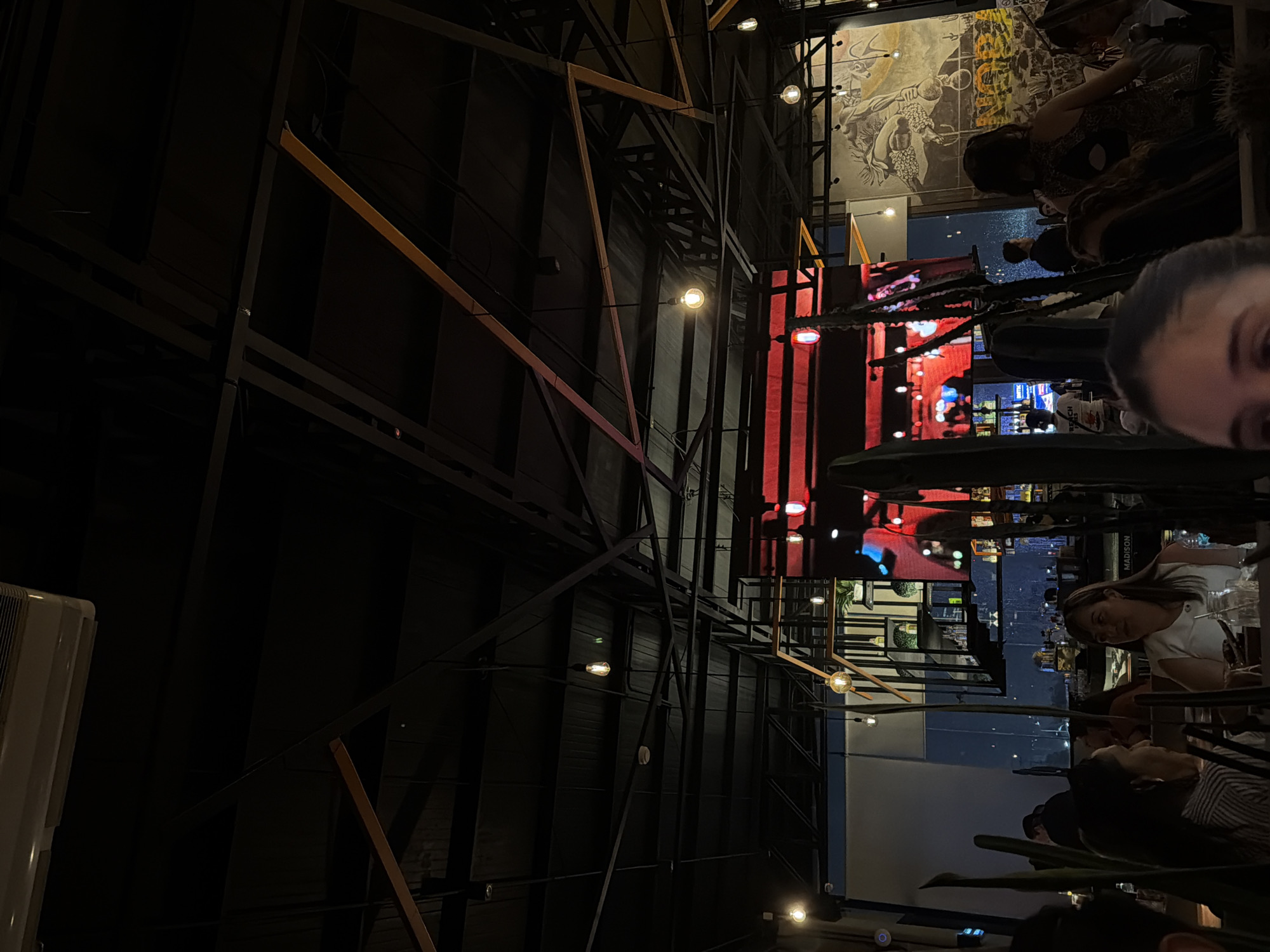 The image shows the interior of a restaurant or bar in Puebla, Mexico, based on the "MADISON" sign visible. Patrons are seated at tables, and a TV screen shows city lights. The interior features exposed structural elements of black metal and brown wood, with decorative lighting. A mural or large graphic is seen on an adjacent wall. A person's face is visible in the lower right corner of the image.