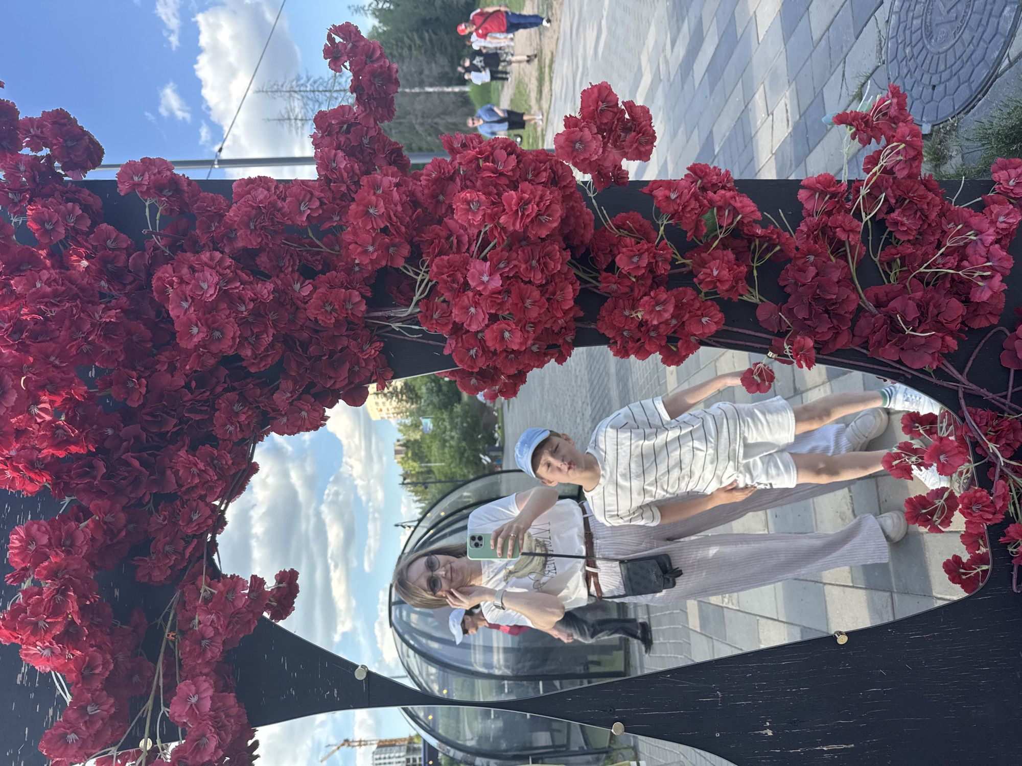 The image captures a woman and a child reflected in a decorative mirror within the Garden of the World (Сад Мира) in Izhevsk, Russia. The mirror features an irregularly shaped dark frame, heavily adorned with clusters of artificial red flowers, consistent with a cherry blossom aesthetic.
In the reflection, a woman with light brown hair, wearing sunglasses, a white t-shirt with a graphic design, light-colored striped pants, and a dark cross-body bag, holds a light green smartphone, appearing to take a photograph or selfie. A metallic watch is visible on her left wrist. Adjacent to her, a young boy wears a light blue baseball cap, a white horizontally striped t-shirt, white shorts, and sneakers. He is making a playful pouting expression and holds a single artificial red flower in his right hand.
Behind the reflected subjects, several transparent, dome-shaped structures, characteristic of geodesic domes, are visible. The immediate ground in the reflection consists of light-colored pavement.
Beyond the mirror's decorative frame, the environment shows an outdoor public space on a clear day with a blue sky and scattered white cumulus clouds. Green foliage and trees are visible in the mid-ground and background. Paved pathways extend into the distance, where several indistinct people are seen walking. On the far left, a construction crane and buildings indicate ongoing urban development, with additional buildings visible in the distant right.