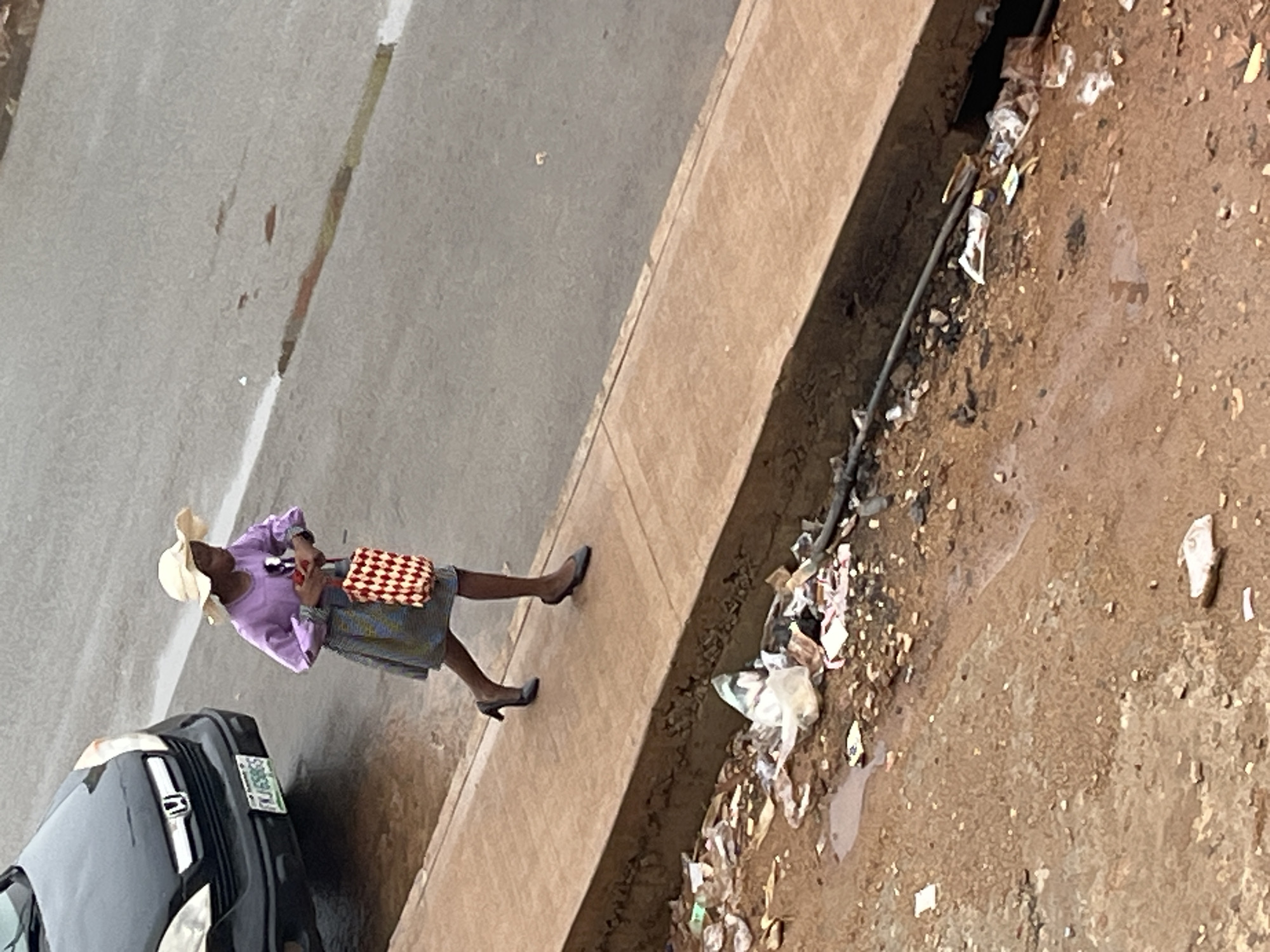 In the city of Jos, Nigeria, a young girl wearing a straw hat and a purple long-sleeved shirt with a colorful patterned skirt walks along a sidewalk. She is carrying a red and white checkered bag. To her left, a black car is parked on the street. The sidewalk is bordered by a ditch filled with debris, including plastic bags and other trash, indicating a poorly maintained urban environment. The ground is wet, suggesting recent rain.
