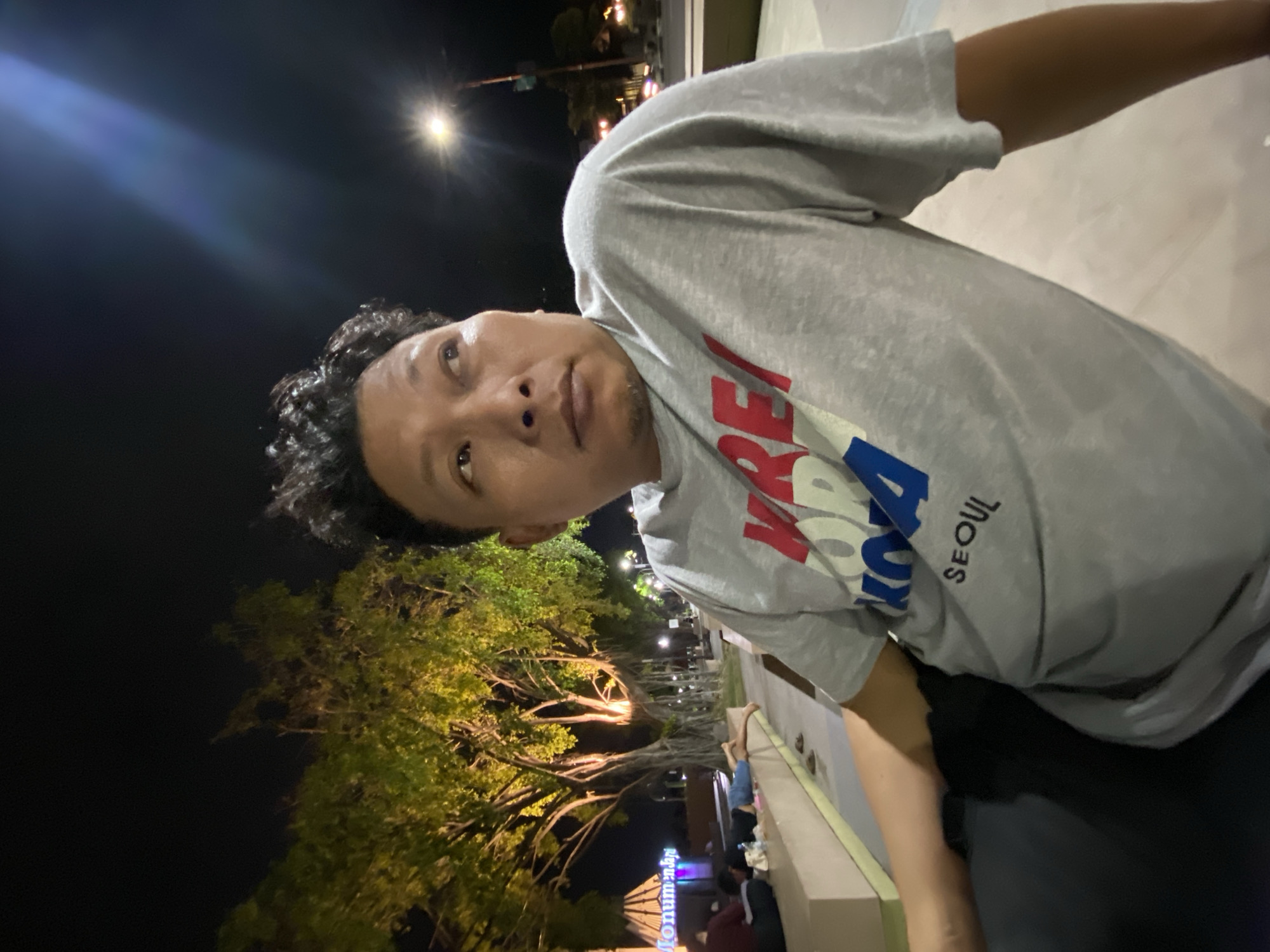 A male individual is captured in a mid-shot photograph taken at night in Sidoarjo, Indonesia, specifically in an area consistent with Monumen Jayandaru. He is of East Asian or Southeast Asian descent, with dark, curly hair and a medium complexion, looking upwards and to his right with a contemplative expression. He wears a light gray short-sleeved t-shirt with the text "KRE" in red, "KO" in white, "REA" in blue, and "SEOUL" in black printed on the front.
The background is an outdoor public space illuminated by various light sources. Large trees with extensive root systems, characteristic of banyan trees, are prominently lit with warm yellow lights on the left, casting a glow on their foliage and trunks. Further left, a large, angular monument structure displays partially visible illuminated text, "Monumen," glowing in blue and purple light. Below the trees, several individuals are visible on a light-colored concrete platform, with at least two lying prone. One individual wears a dark top and light pants, while another wears a red top. A pair of sandals is visible on the platform. On the far right, a bright streetlight casts a prominent lens flare, and distant vehicle lights are faintly visible on a paved surface. The overall scene suggests a vibrant public park or monument area at night.