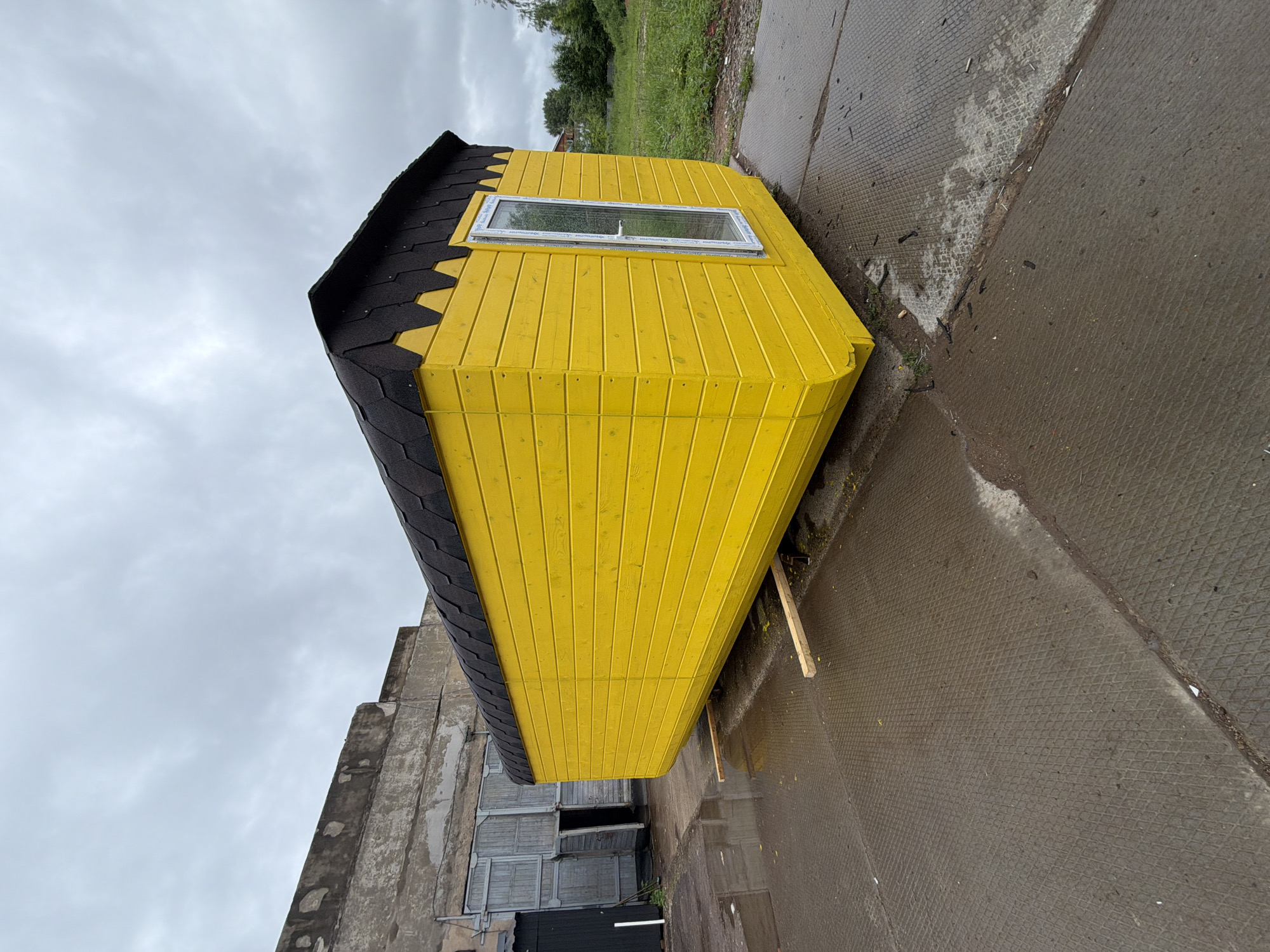 The image depicts an outdoor industrial or storage yard in Votkinsk, Russia, under an overcast sky. The primary subject is an elongated rectangular structure, likely a portable cabin or shed, painted in a vibrant yellow. Its exterior is clad in horizontal wooden planks. The roof is covered with dark brown or black scalloped asphalt shingles, featuring a noticeable overhang. A single, tall, narrow, white-framed window or door with a visible handle is situated on its right-facing side. The structure is elevated slightly off the wet ground by two light-colored wooden planks or runners.
The ground is a textured concrete or paved surface, displaying a distinct diamond or grid pattern. It is thoroughly wet with standing water and reflections, indicating recent precipitation. Scattered small dark debris, possibly wood fragments or construction waste, lie on the surface, particularly near the base of the yellow structure and along a visible seam in the pavement.
In the mid-ground, to the left of the yellow structure, stands a dilapidated, multi-story building with weathered, crumbling concrete or stucco walls. Two large, faded gray wooden double doors are visible on this building; one is partially open, revealing a dark interior. A smaller, darker doorway is also seen to the far left. A black object, possibly a bin or lean-to, is partially visible on the extreme left, with a white vertical pole adjacent to it.
To the right of the yellow structure and in the distant background, a patch of overgrown green grass, weeds, and various trees and bushes marks the edge of the paved area, with faint outlines of distant structures visible through the foliage. The sky is uniformly light gray with dense cloud cover. No individuals or observable actions are present within the frame.