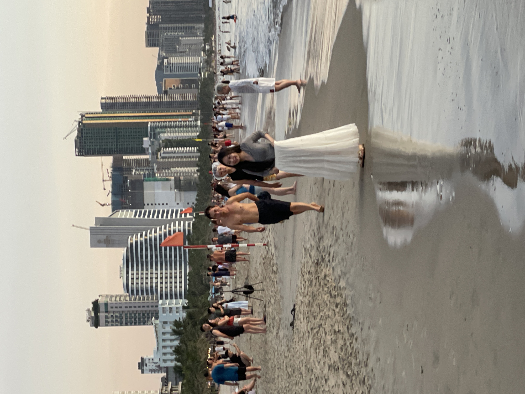 This image depicts a crowded beach scene in the city of Đà Nẵng, Vietnam, with a backdrop of modern skyscrapers. The foreground features a sandy beach with several people walking, some near the water's edge. A man with a bare torso and dark shorts is walking towards the camera, while a woman in a white skirt and grey top is standing to his left, looking away. In the background, numerous high-rise buildings, some under construction, line the coastline. The ocean is visible with small waves, and several people are in or near the water. Red flags are planted in the sand. The sky is a soft, hazy orange, suggesting either dawn or dusk.