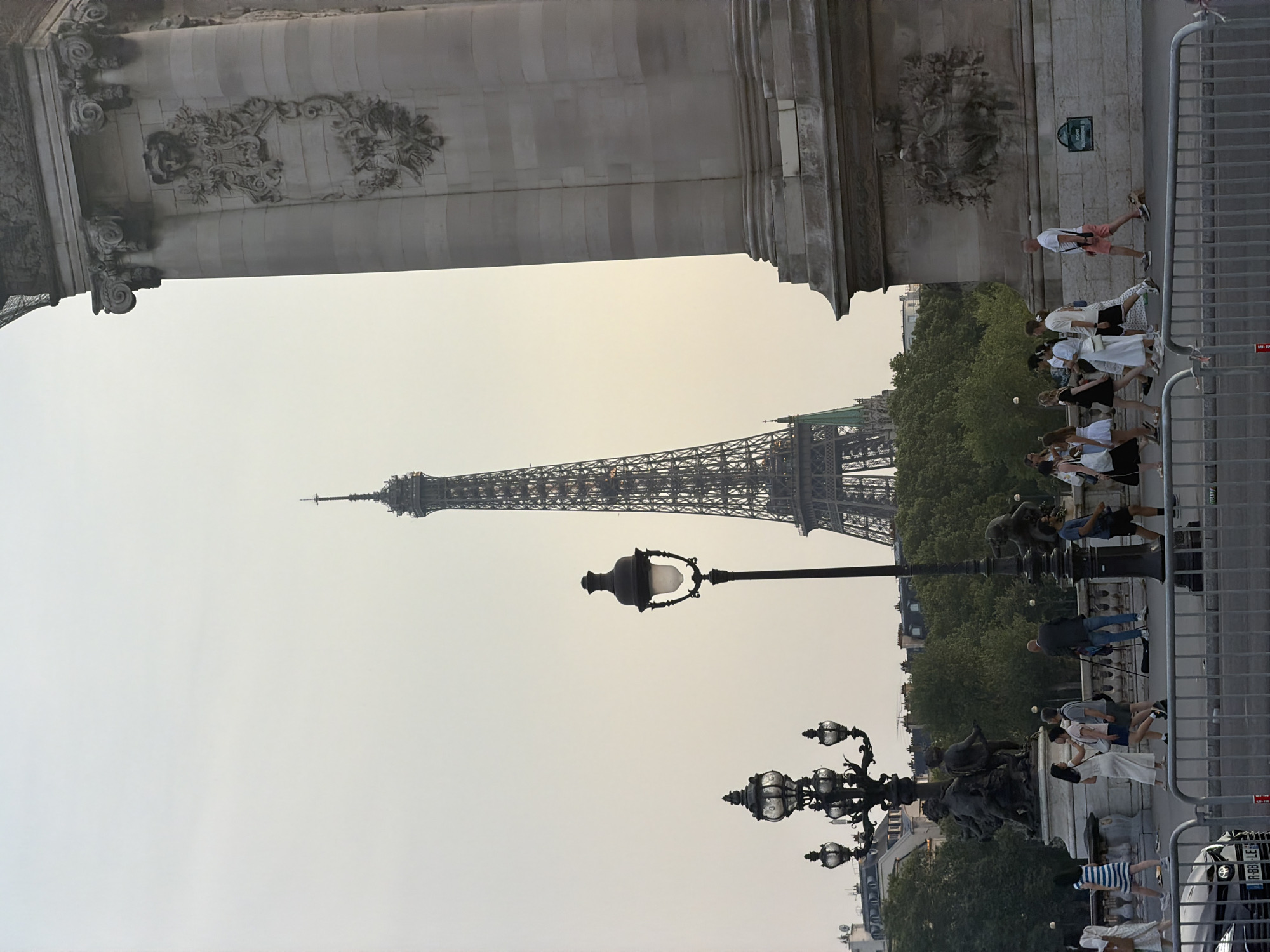 The image captures a view of the Eiffel Tower in Paris, France, from a pedestrian bridge, likely the Pont Alexandre III, given the ornate architectural elements visible on the right. The Eiffel Tower is prominently featured in the background, appearing tall and dark against a hazy sky. In the foreground, a street lamp with multiple globes stands to the left, and another single, larger street lamp is positioned more centrally. A group of pedestrians is walking across the bridge from left to right. They are casually dressed, suggesting a daytime scene. The bridge itself has a metallic barrier at the bottom edge of the frame, and behind the pedestrians, there are trees and buildings. The overall atmosphere is somewhat muted due to the hazy sky.