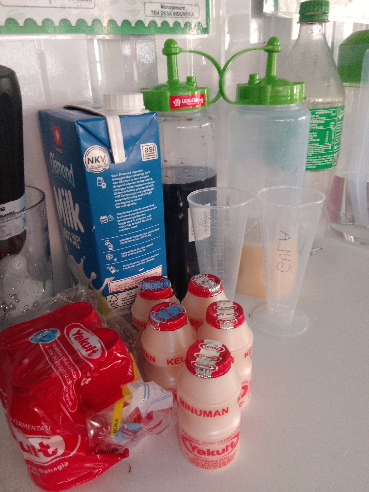 The image shows a collection of drinks and bottles on a white surface. In the foreground, there is a bright red plastic bag with the "Yakult" logo and text that appears to be in Indonesian, indicating "Fermentasi" and "Bahagia". Several small bottles of Yakult with red caps are arranged around the bag. Behind the Yakult, there is a blue carton of "Diamond Milk" with Indonesian text. To the right of the milk carton, there are clear plastic cups and larger bottles with green lids. One of the cups has the name "AJI" written on it in black marker. In the background, there are more bottles and a white, textured wall with some green decorative elements that might be part of a sign or advertisement, also with Indonesian text. The lighting suggests an indoor environment. The items are likely on a table or counter in a shop or a home kitchen, possibly in Trenggalek, Indonesia, given the presence of Indonesian language products.