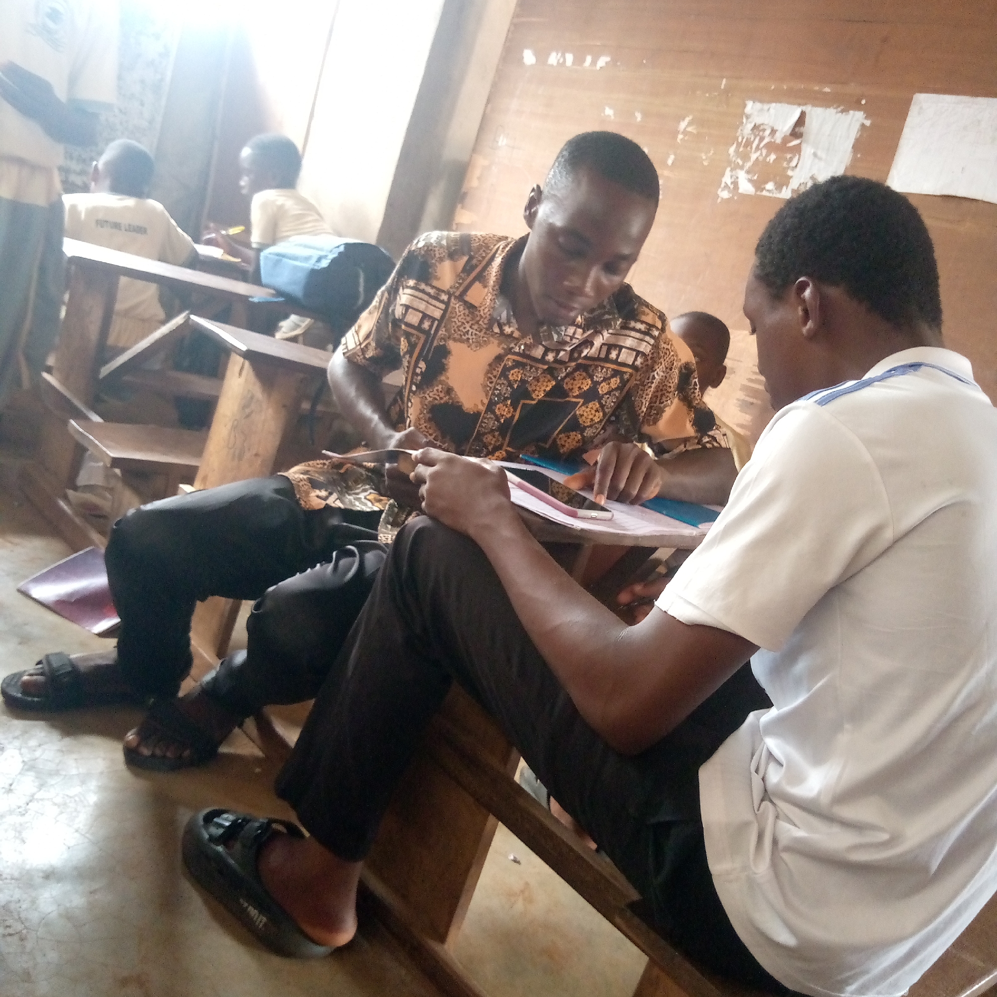 The image depicts two young men sitting at a wooden school desk in what appears to be a classroom setting in Ukwu-Abwa, Nigeria. The man on the left wears a patterned shirt and dark trousers, while the man on the right wears a white shirt with blue trim and dark trousers. They are both looking at a document on the desk, possibly working together on an assignment. Other students are visible in the background, seated at similar desks, with one wearing a shirt that says "FUTURE LEADER." The classroom environment appears modest, with a wooden floor and walls. The lighting is bright, suggesting daylight.