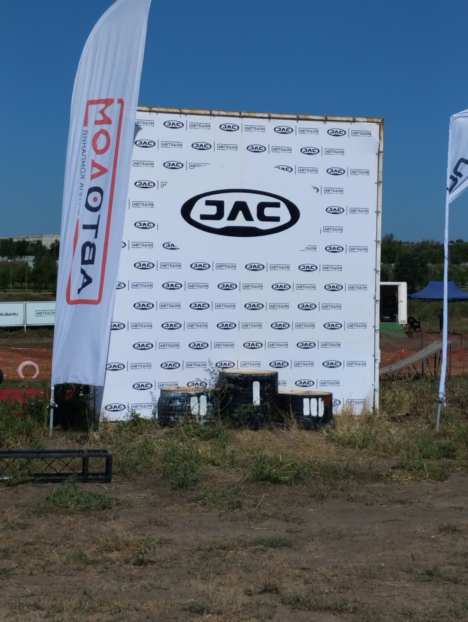 This media file captures an outdoor scene, likely at a promotional event or race, in Kostanay, Kazakhstan. The primary focus is a large white banner with the JAC logo prominently displayed, along with numerous smaller repeated logos and the text "АВТОДОМ" (Avtodom), indicating a car dealership or service. The banner is positioned outdoors, with a clear blue sky suggesting a sunny day. In the foreground, dry grass and dirt ground are visible, along with some industrial-looking barrels, possibly used as obstacles or decorations. To the left of the banner, a tall white flag with red lettering, "ΜΟΛΟΤΟA" (Molotova), also appears to be a promotional item. In the background, glimpses of trees, buildings, and a blue tent can be seen, hinting at a more established event space or an adjacent area. There are no discernible people or animals in the frame. The overall impression is one of an open-air event showcasing automotive brands, possibly related to motorsports or a car exhibition.