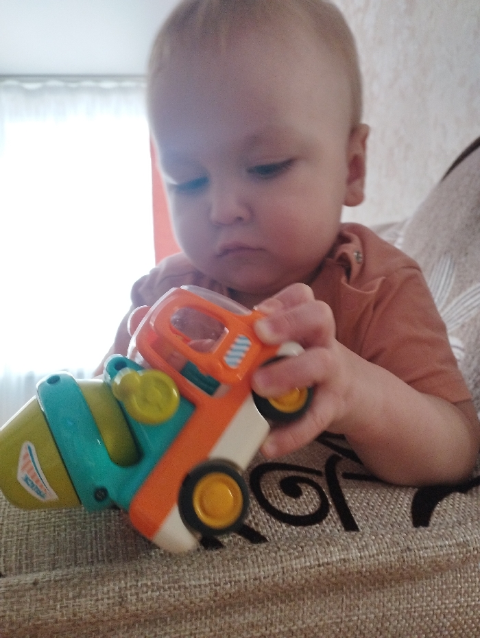 A baby sits on a tan couch, holding a colorful toy truck. The truck is mostly orange and teal with yellow accents. The baby is fair-skinned with light hair, wearing a short-sleeved orange shirt. They are looking down at the truck with a serious expression. The background includes a window with a sheer white curtain. The couch has a decorative pattern.