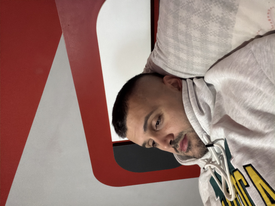 An indoor scene shows a male subject reclining. The subject has short dark hair with faded sides and short facial hair consisting of a beard and mustache. He wears a light grey hooded sweatshirt with a yellow and green design on the chest, and white drawstrings are visible at the neckline. His head is resting on a light grey patterned pillow. The subject's eyes are directed downwards and slightly to his left. The background features a distinctive red and white structure with curved and angled panels, consistent with a modern bed frame or wall paneling. A dark rectangular area is visible through an opening in the red panel on the left. The precise location within Opanets, Bulgaria, is not discernible from the visual information provided.