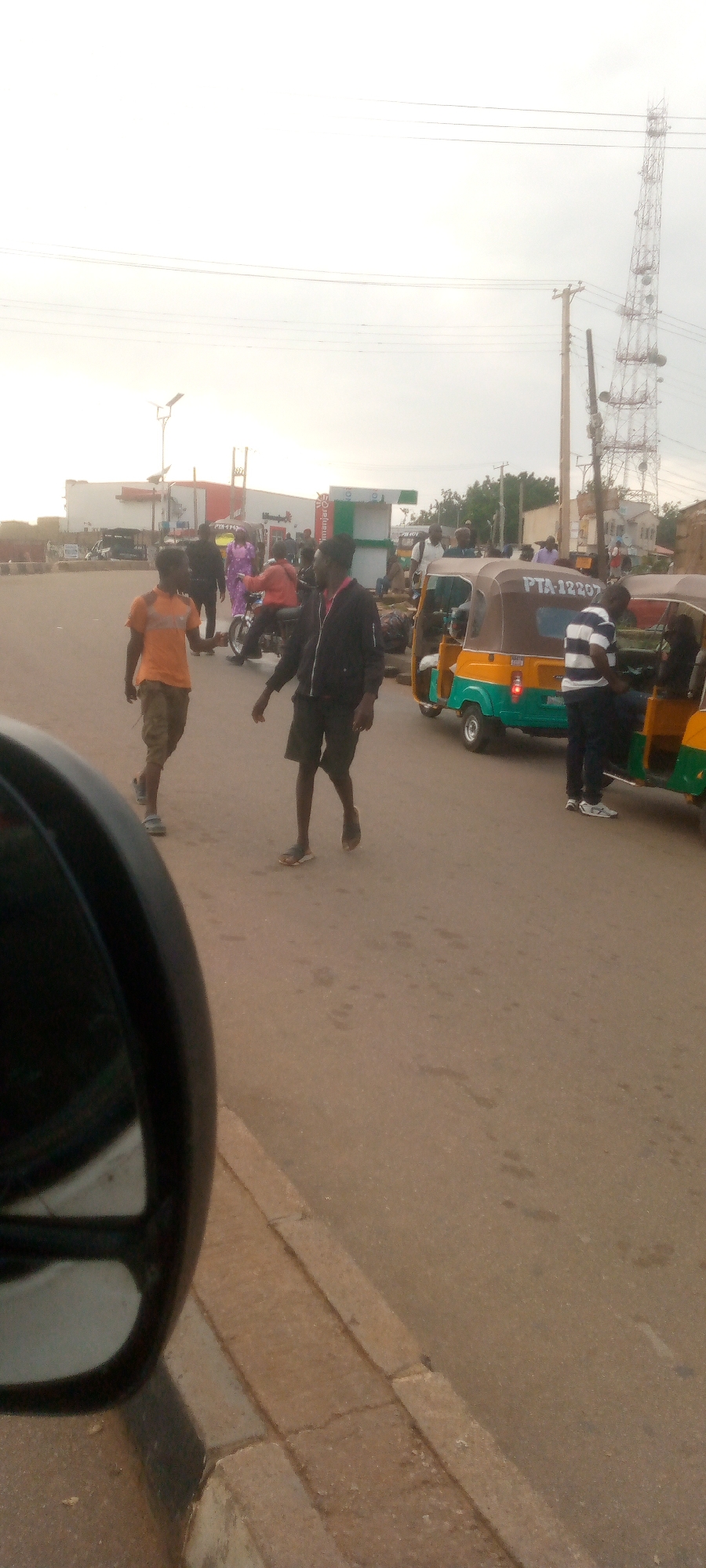 The image depicts an outdoor scene on a dusty, paved road, likely in Gura Top, Nigeria. The time of day appears to be late afternoon or early evening, judging by the soft lighting. In the foreground, the reflection of a black side mirror from a vehicle is visible on the left.
The main focus of the image is a street intersection with several people and vehicles. Two young men are walking on the road; one in an orange t-shirt and khaki shorts, the other in a black jacket and dark shorts. Behind them, a woman in a vibrant purple dress is visible.
Further into the scene, a motorcycle is being ridden by a person in an orange shirt. Several auto-rickshaws, also known as 'Keke Napep' or 'Marwa', are present. One yellow and green auto-rickshaw has the license plate "PTA-12207" clearly visible. A man in a blue and white striped shirt is standing next to this auto-rickshaw.
In the background, a large telecommunications tower with multiple platforms and antennas dominates the skyline. Other structures, possibly shops or market stalls, are lined up along the side of the road, and some vehicles are parked. The environment is characterized by the general hustle and bustle of a busy urban street.