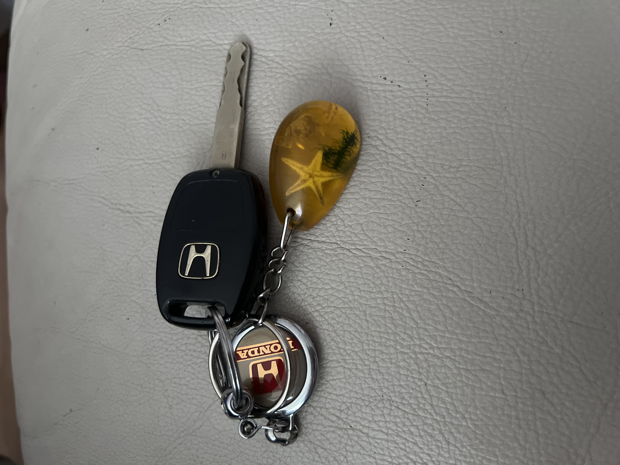 A Honda car key and a keychain with a Honda logo and a transparent resin charm containing a starfish and seaweed are placed on a light-colored textured surface, possibly leather. The keychain is attached to the key with a silver ring and chain. The image is taken from a top-down perspective. The scene is not identifiable as being in Vilnius, Lithuania.