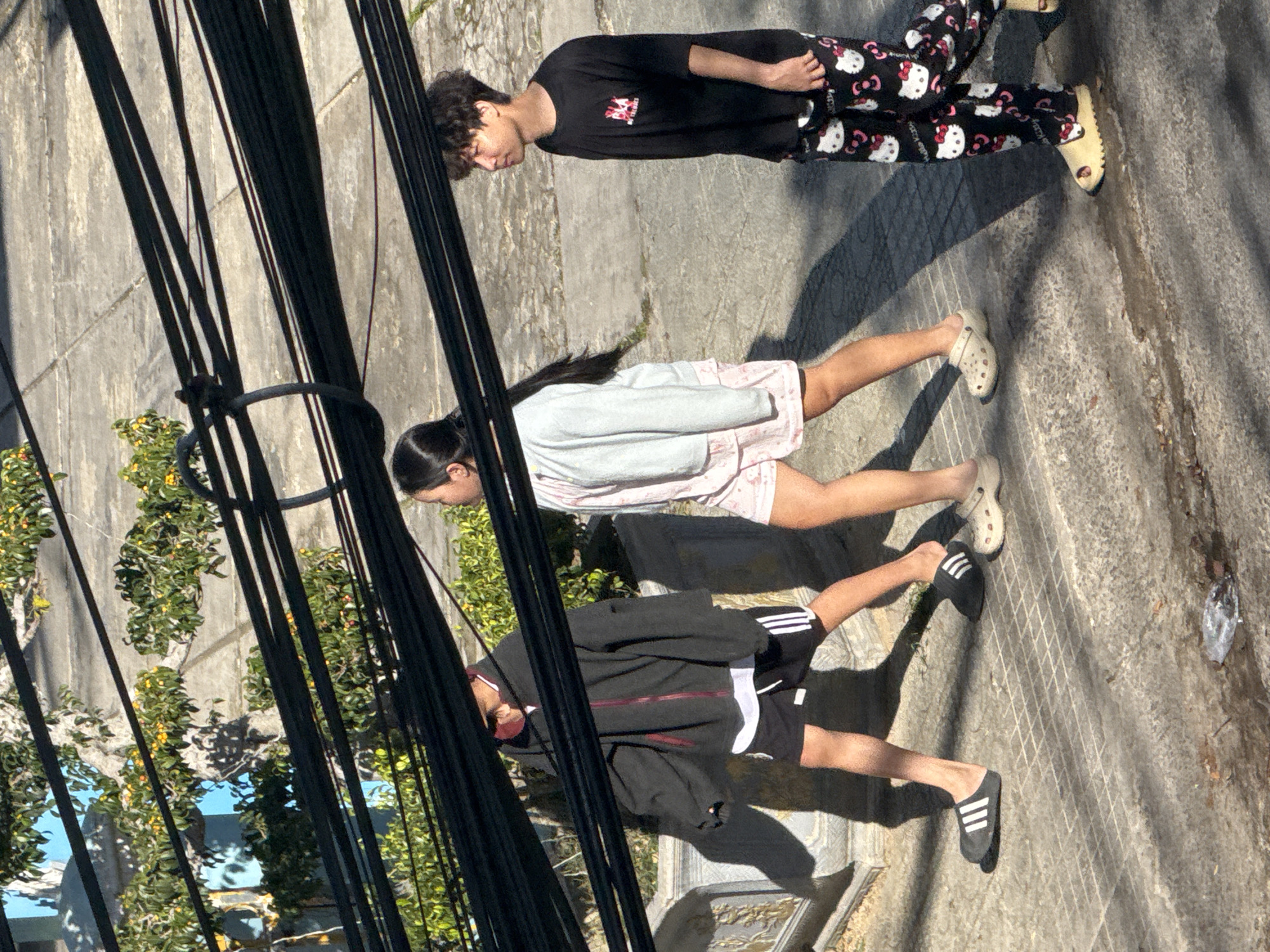 The image shows three young people walking on a paved sidewalk in Da Lat, Vietnam. The weather appears sunny with shadows indicating a midday sun. Overhead, a dense network of black electrical wires spans across the frame.
On the left, a person wearing a black zip-up hoodie, black shorts with white stripes, and black Adidas slides walks with their head down. A portion of a white t-shirt is visible under the hoodie.
In the middle, a person in a light gray zip-up hoodie and pink shorts with a white pattern walks alongside. They are wearing beige Crocs. Their dark hair is pulled back in a ponytail.
On the right, a person with curly dark hair wears a black t-shirt with a graphic print and black pants adorned with a repeating pattern of Hello Kitty faces and bows. They are wearing cream-colored platform slides. Their gaze is directed to the side.
The background consists of a textured gray pavement, a low stone wall with ornate carvings, and some greenery with yellow flowers. The environment suggests an urban or semi-urban setting.