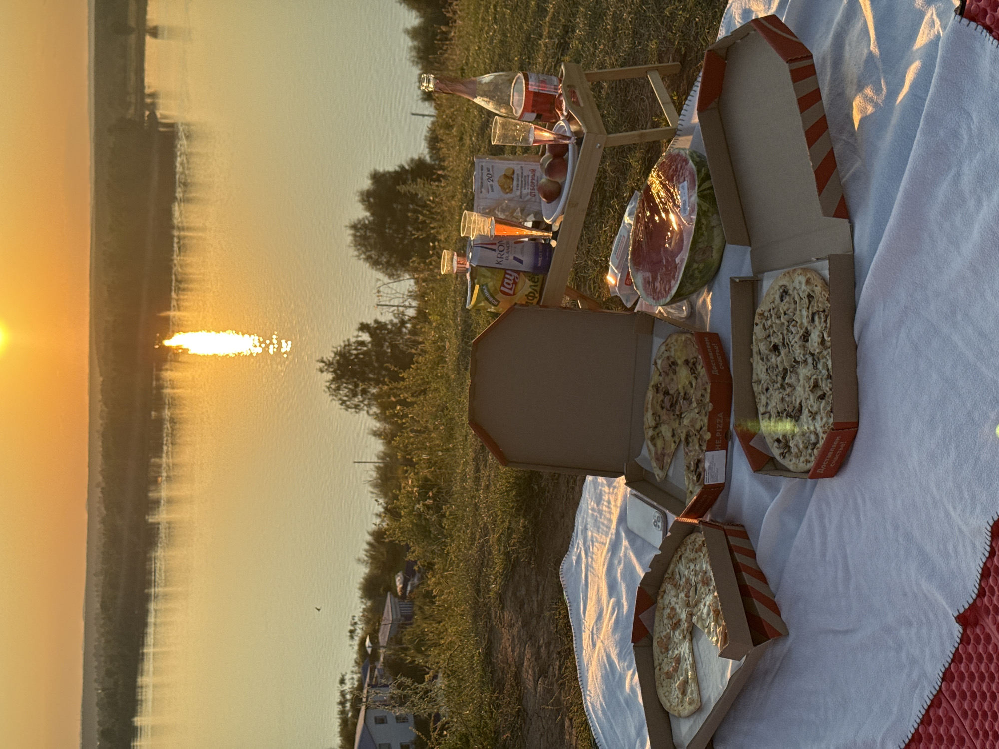 This is a forensic, matter-of-fact description of a picnic scene set against the backdrop of a sunset over a body of water, likely the Volga River, near the city of Olkhovka, Russia.
The scene is set on a grassy slope overlooking a wide river. The sky is ablaze with the warm hues of sunset, casting a golden glow on the water. A sunlit reflection stretches across the river's surface. In the distance, the opposite bank is lined with trees and a hazy horizon. On the near bank, some buildings are visible, suggesting proximity to a settlement.
In the foreground, a white picnic blanket is spread out. On the blanket, there are several pizza boxes. One box is open, revealing a pizza with a visible crust and toppings. Another open box contains a pizza with a single slice missing. A third pizza box is also open and shows a pizza with mushroom toppings. A half of a watermelon, wrapped in plastic, is positioned near the pizzas.
A small portable wooden table is set up. On the table, there are several items: a bottle of pink sparkling wine, a bottle of what appears to be mineral water, three clear plastic cups with pink liquid, two bags of potato chips, and a small plate with three pieces of fruit, likely peaches. A white mobile phone is also visible on the blanket.
The overall scene depicts a leisurely outdoor meal during a sunset. There are no people visible in the image. The environment is natural, with grass and trees, and the presence of the river and distant buildings indicates a semi-rural or edge-of-town setting.