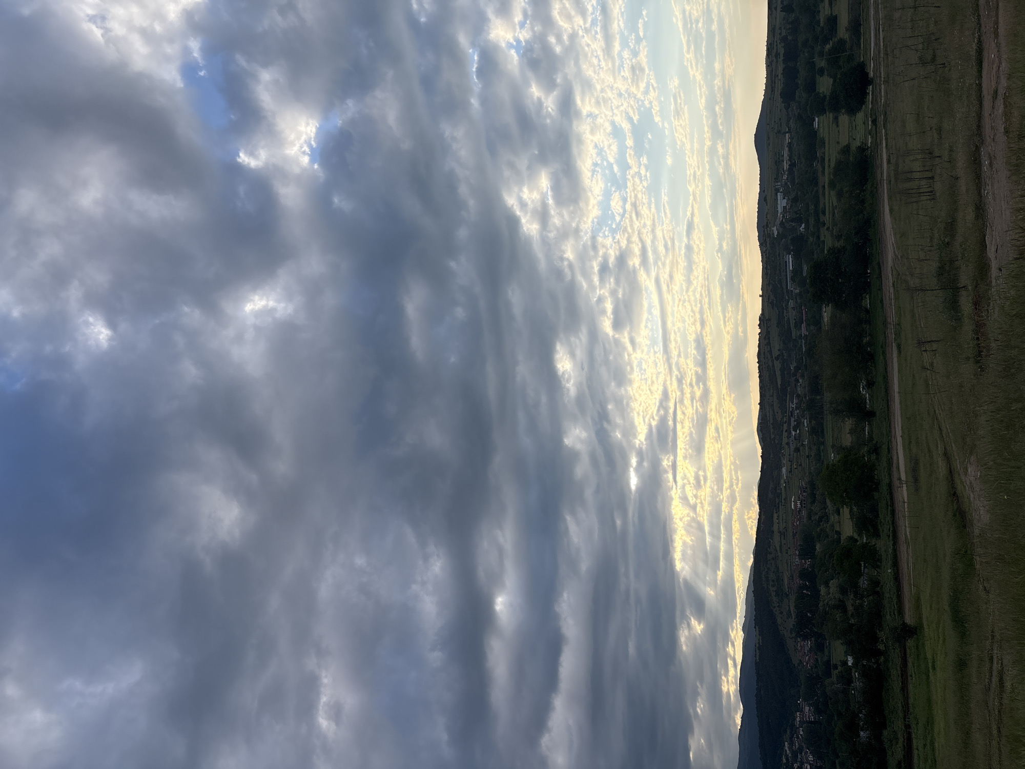 The image shows a landscape view in Strelcha, Bulgaria. The left side of the frame is dominated by dark, heavy clouds in the sky. The clouds exhibit varying shades of gray and white. On the right side of the image, there is land, partially covered in vegetation and showing signs of human development. Some buildings and trees are visible on the slope of a hill. The land is illuminated by light that filters through the clouds.