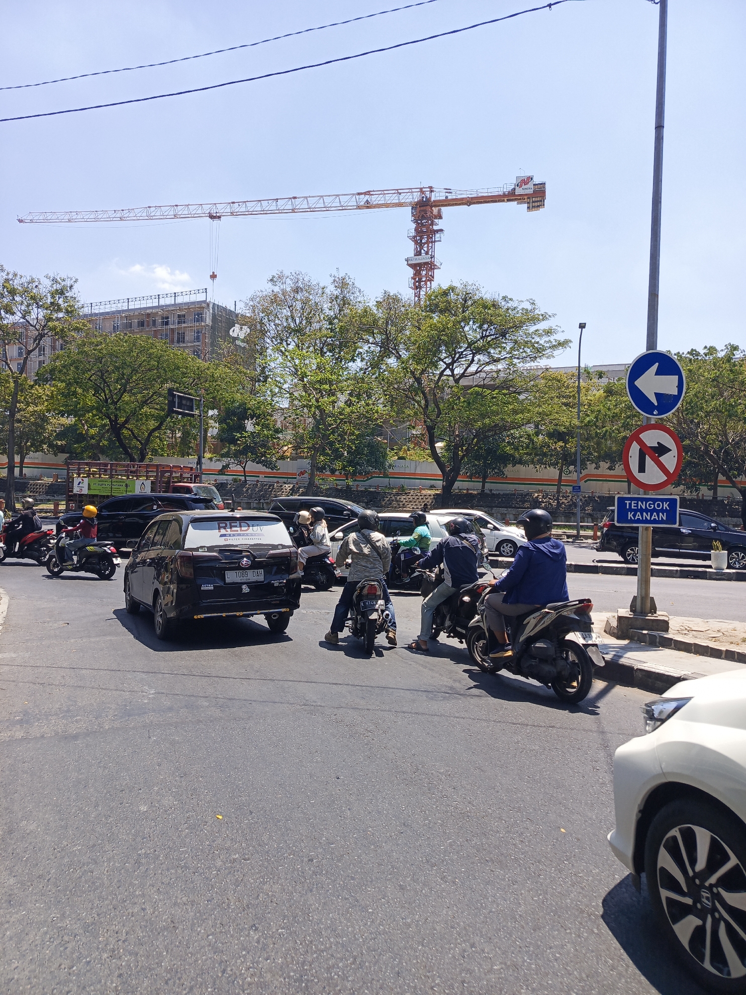 This image captures a street scene in Surabaya, Indonesia, under a clear sky. In the background, a tall construction crane is visible above a multi-story building under construction, with scaffolding and unfinished sections. Trees with lush green foliage line the street in front of the building.
The foreground is dominated by a road with moderate traffic. Several motorcycles are in motion or stopped, carrying one or two riders wearing helmets. A black car is in the left lane, and a white car is partially visible on the right side of the frame. Traffic signs are present, including a blue circular sign with a white arrow pointing left, a red and white circular sign with a diagonal line across an arrow pointing right, and a blue rectangular sign with white text reading "TENGOK KANAN" which translates to "LOOK RIGHT." A lamppost stands to the right of the traffic signs. The road surface is asphalt, showing wear and markings. The overall impression is a busy urban intersection in a tropical climate, indicated by the bright sunlight and greenery.
