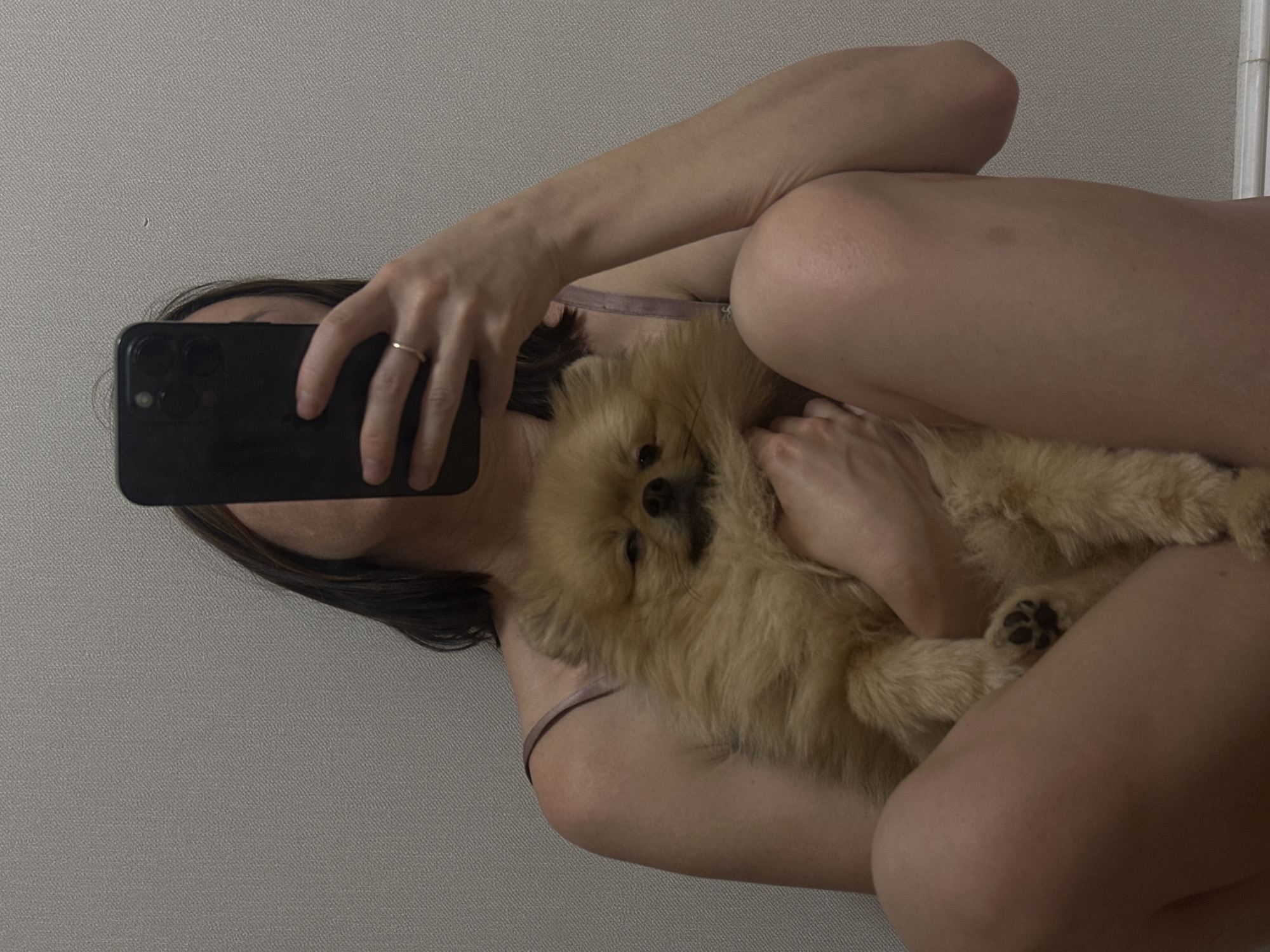 A medium shot captured from a low angle shows a person seated with their legs crossed, holding a small, fluffy, light brown Pomeranian dog. The person is holding a black smartphone up to take the photo, obscuring their face. A thin gold ring is visible on the ring finger of the hand holding the phone. The person is wearing a light-colored camisole. The background is a plain, textured light gray wall. The lighting is soft and appears to be artificial. This image was taken indoors.