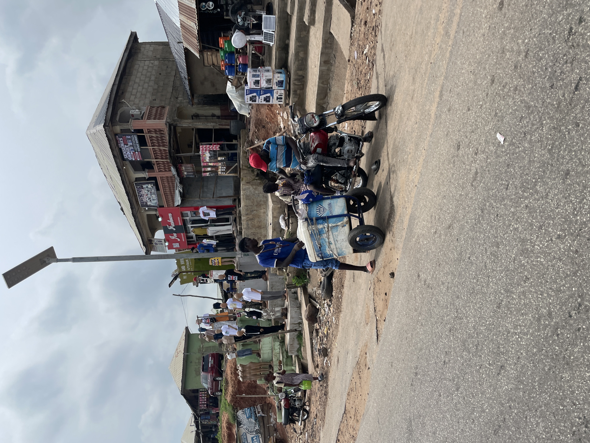 In Lokoja, Nigeria, a street scene unfolds with a man pushing a cart with a blue and white cooler, labeled "Fresco Milk." He is dressed in a blue t-shirt and denim shorts. Seated on a red motorcycle beside him is another individual wearing a striped shirt and a red cap, also in denim. The motorcycle is parked on a dirt road adjacent to a paved area. Behind them, a cluster of mannequins is displayed on raised steps in front of shops. These mannequins are dressed in various casual outfits. The shops appear to be small, local businesses, with signs visible, including "Joe Fashion" and "Sensema's Gadget Store." One building has a multi-story structure with a balcony. A red SUV is parked on the left side of the scene. The environment suggests an outdoor market or commercial area in a town or city, characterized by a mix of shops, display areas, and pedestrian activity. The sky is overcast.