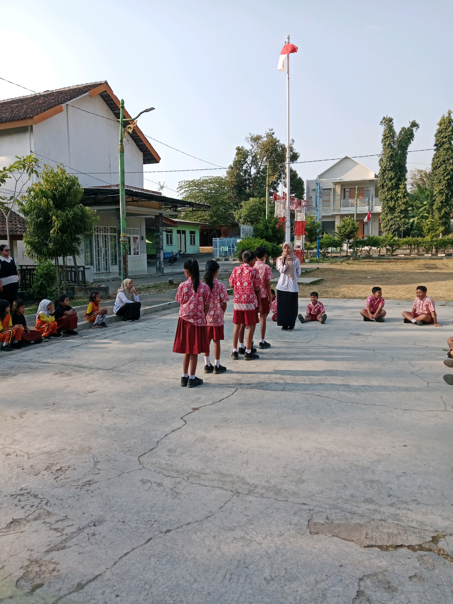 The image depicts an outdoor scene at what appears to be a school in Salatiga, Indonesia, likely during a flag ceremony or morning assembly. In the background, a two-story building with a light-colored facade and a reddish-brown tiled roof is visible, along with other school structures and trees. A flag pole with the Indonesian flag (red and white) is prominently featured.
In the foreground, a group of children dressed in school uniforms are gathered. Several young girls in matching red and white patterned shirts and red skirts are standing, facing away from the camera, while a teacher or instructor in a light-colored shirt and dark skirt stands before them, holding a microphone. To the left, a group of children, some in similar uniforms and others in different colored attire, are seated on the ground or a low wall, watching the proceedings. Some of these seated children wear orange tops and dark bottoms or skirts. To the right, a few boys in red and white patterned shirts and dark shorts are also seated on the ground. The ground is paved with concrete, showing cracks and some wear. The overall lighting suggests it is daytime, likely in the morning, given the presence of a flag ceremony.