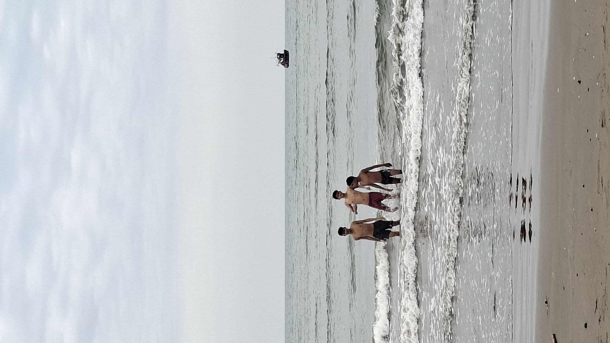 The image shows three shirtless boys wading in the ocean near the shore, with a small fishing boat in the distance. The sky is overcast and the water is choppy with white waves. The boys are standing in shallow water, and the one in the middle appears to be splashing. The beach in the foreground is sandy. Based on the presence of fishing boats and the coastal environment, this scene is likely from a beach in Lhokseumawe, Indonesia.