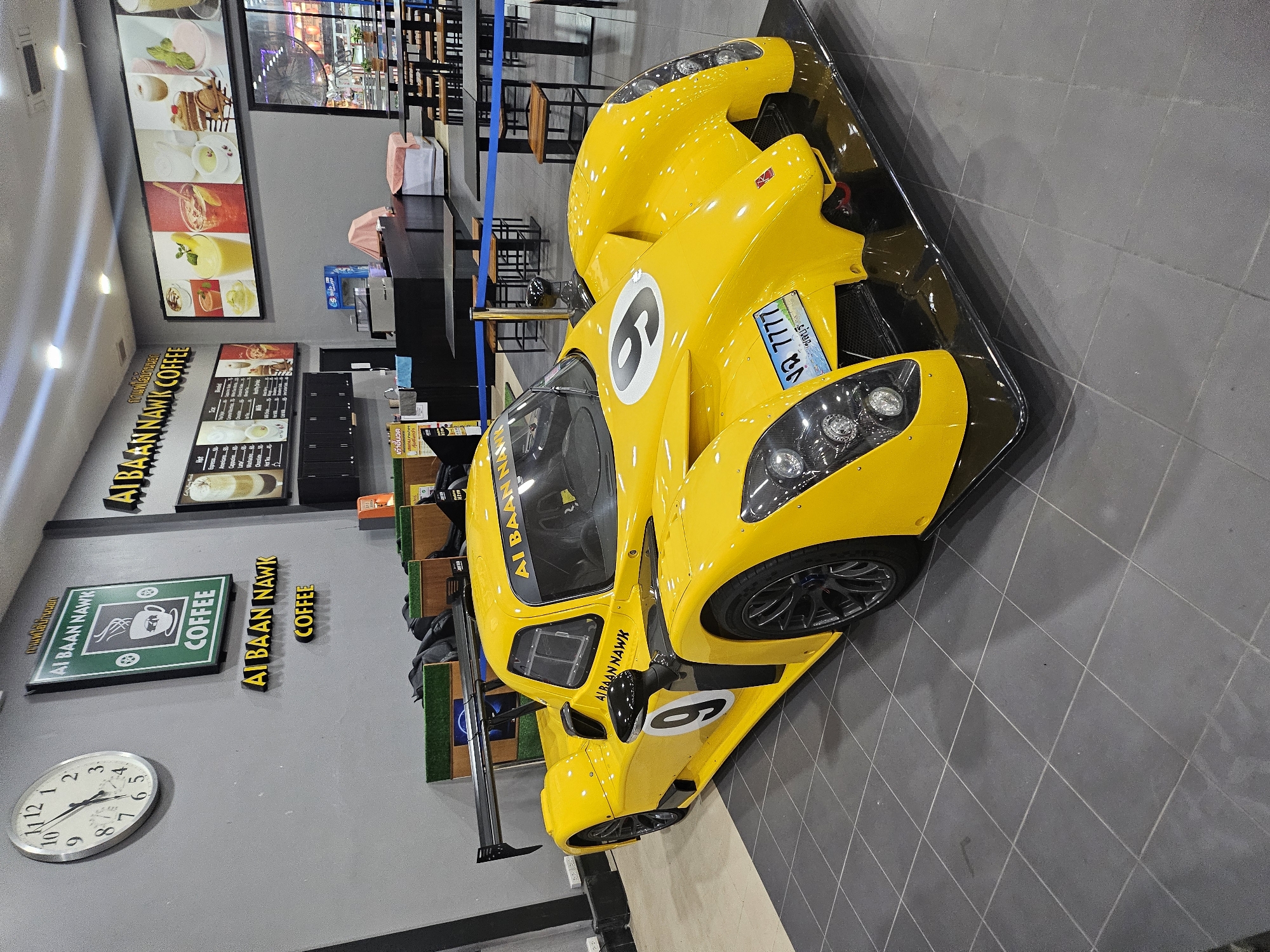 In Na Chom Thian, Thailand, a bright yellow racing car is displayed indoors, positioned on a tiled floor. The car, a prototype with a prominent rear spoiler and aerodynamic bodywork, is the central focus. A large number '9' is visible on the side of the car, and another '9' is on the hood. The car has a license plate with Thai characters and the number '7777'.
Behind the car, the environment appears to be a cafe or restaurant. A large circular clock with Roman numerals is mounted on a grey wall to the left. Above the clock, a green sign with white lettering reads "AI BAAN NAWK COFFEE". Below this, a similar sign in yellow 3D lettering says "AI BAAN NAWK COFFEE". Further to the right, a long panoramic image displays various colorful drinks and desserts. A menu board with Thai text is visible on the wall to the left of the panoramic image. In the background, several tables and chairs are set up, suggesting a dining or seating area. The overall lighting is artificial, with ceiling lights illuminating the space. There are no people visible in the image.