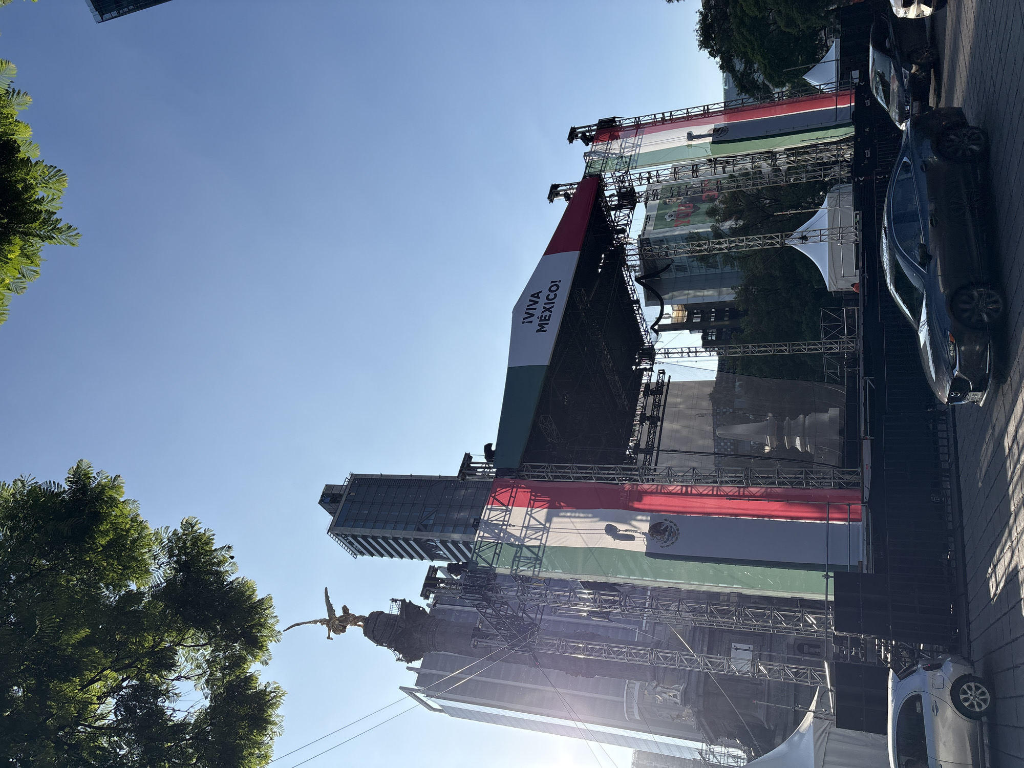 This image depicts a large outdoor stage draped with the Mexican flag and banners, set up in a public space. The Mexican flag is prominently displayed in vertical green, white, and red stripes, with the national coat of arms visible on the white stripe. A large banner with the text "¡VIVA MÉXICO!" is also visible on the stage. The stage is constructed with scaffolding and appears to be part of a celebration or event, likely related to Mexican Independence Day, given the prominent display of national symbols.
In the background, modern skyscrapers rise above the stage, suggesting an urban setting. A golden statue of an angel, likely the Angel of Independence monument, is visible on the left, further confirming the location as Mexico City. Trees with lush green foliage frame the left side of the image, providing a natural element to the urban landscape.
Several cars are parked along the side of the street next to the stage, indicating that the event is accessible by road. The sky is a clear, bright blue, suggesting it is daytime with good weather, likely a sunny afternoon. The overall atmosphere conveyed is festive and patriotic, celebrating Mexican culture and national pride. The image is oriented vertically, with the sky appearing at the top and the ground at the bottom.