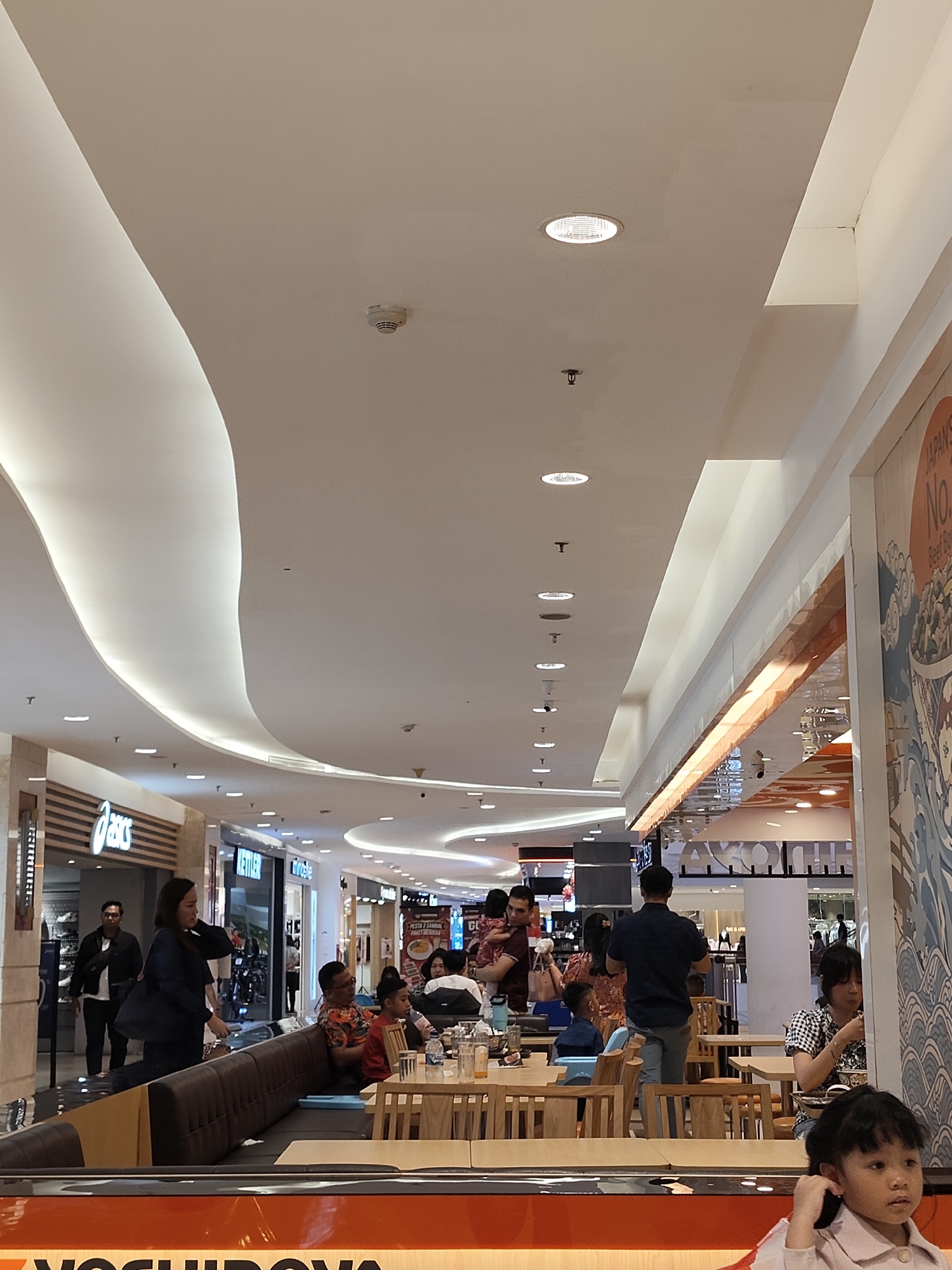 The image shows an interior view of a brightly lit shopping mall, likely in the City of Medan, Indonesia, based on context provided elsewhere. The scene is dominated by a high, modern ceiling with curved, illuminated sections and recessed lighting. In the foreground, a girl with dark hair is looking to her right, partially visible from the chest up, wearing a light-colored top. She appears to be seated at a table. Behind her, patrons are seated at tables, some eating and some conversing. Several individuals are visible, including a man in a dark blue shirt and grey pants standing and facing away from the camera, and a woman in a dark jacket walking past. Storefronts are visible in the background, including "Asics" and "Kettler," indicating a retail environment. The overall atmosphere is that of a busy commercial space, likely a food court or dining area within the mall.