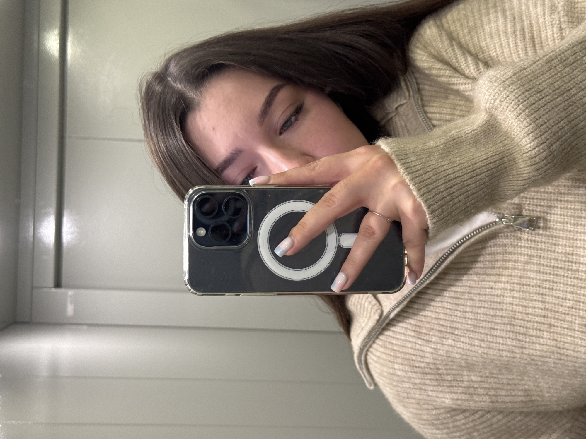 A young woman with long, dark brown hair is taking a selfie in a dimly lit room with beige walls. She is wearing a light beige, ribbed knit sweater with a zippered collar. Her right hand is holding a dark-colored smartphone in front of her face, partially obscuring it. The phone has a clear case with a silver magnetic ring visible on the back. She has manicured nails with a French tip design that fades into light blue. A silver ring is visible on her ring finger. The background appears to be an elevator interior with paneled walls. The lighting suggests an indoor, possibly urban, environment.