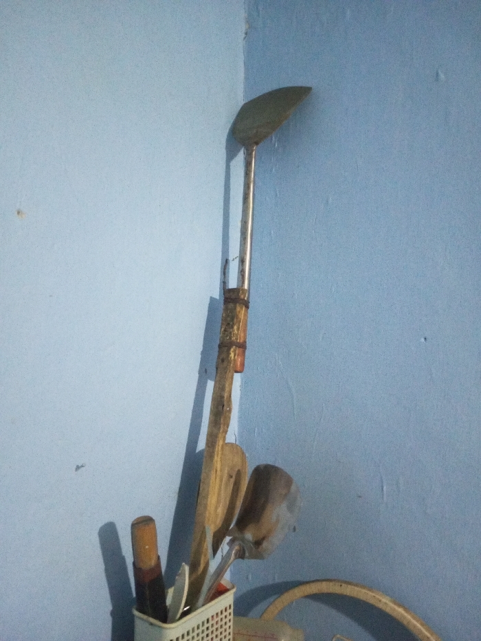 The scene is an indoor corner in Ciherang, Indonesia, featuring two intersecting walls painted a light blue. Various kitchen utensils are leaning against the corner and contained within a holder. Dominating the upper portion is a long-handled metal spatula with a curved, flat head, its metallic handle extending downwards. Parallel to it, a wooden spatula or paddle leans against the wall, exhibiting a darker, possibly reinforced section on its handle. In the lower left, a white, rectangular, perforated plastic holder contains several items, including a knife with a dark handle and at least one other metallic utensil. A light-colored, curved object, possibly part of another container or tool, is partially visible in the lower right foreground. Shadows are cast along the left wall, indicating a light source from the right. No individuals are present.