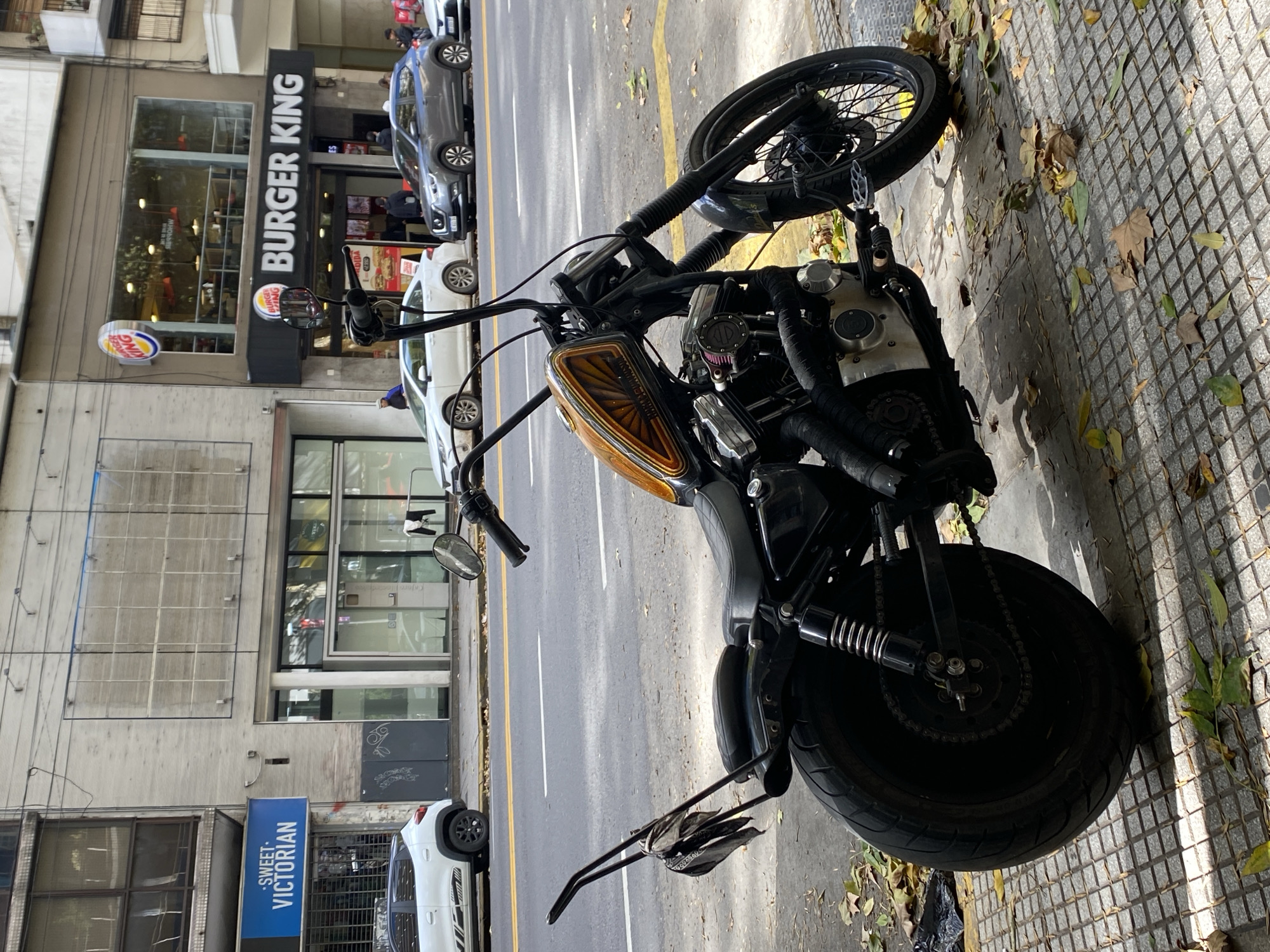 A black motorcycle is parked on a sidewalk in Buenos Aires, Argentina. The motorcycle has a black frame, front and rear tires, and a brown fuel tank. A Burger King restaurant is visible in the background, as well as a cafe named "Sweet Victorian". There are cars parked along the street, and pedestrians can be seen in the background. The sidewalk is paved and covered with fallen leaves. The street has yellow traffic lines.