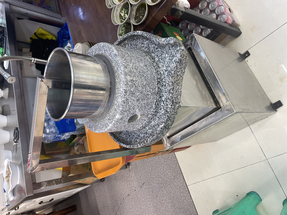 The image shows a stone mill in a food stall in Chongqing, China, located at coordinates 29.58° N, 106.53° E. The mill is made of a circular gray stone base with a smaller, circular metal bowl on top, filled with liquid. The base is surrounded by a stainless-steel basin, which is connected to a metal frame with a metal pipe extending to the top of the bowl. The mill is situated on a tiled floor, next to a stainless-steel counter with various items on it, including a yellow and orange plastic container. Several small white bowls filled with chopped green vegetables can be seen on a wooden table beside the mill, suggesting the preparation of food. A few cans of beverage are visible in the lower right corner of the image. The image was taken at eye level, facing the mill.