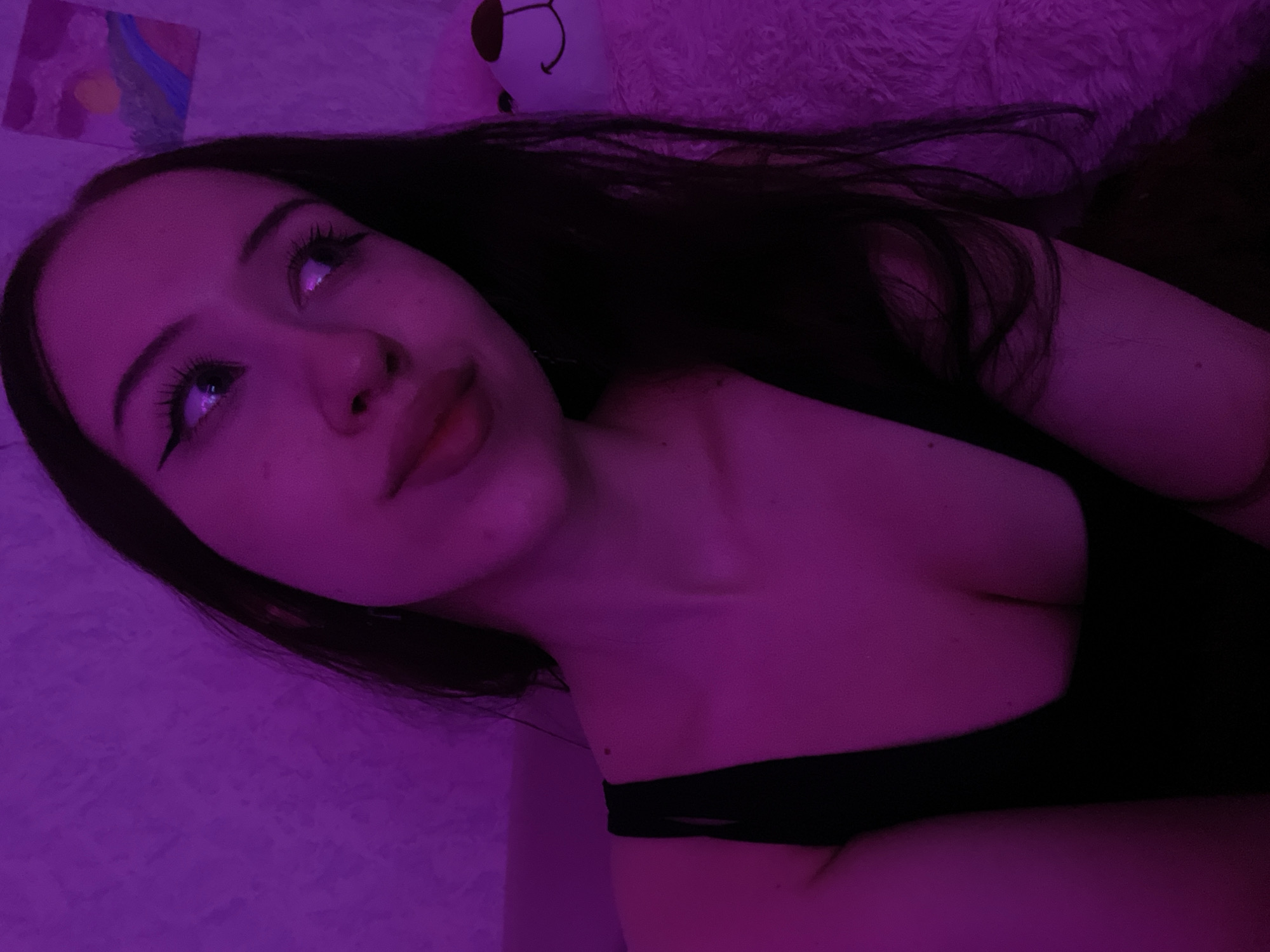 A young woman with long, dark hair and prominent eye makeup is captured in a low-angle, close-up shot, illuminated by purple lighting. She is wearing a black, sleeveless top. Her gaze is directed upwards and to her right. A stuffed bear with a smiley face is partially visible to her right, and a small painting hangs on the wall behind her. The background appears to be a textured wall. The location is Votkinsk, Russia.