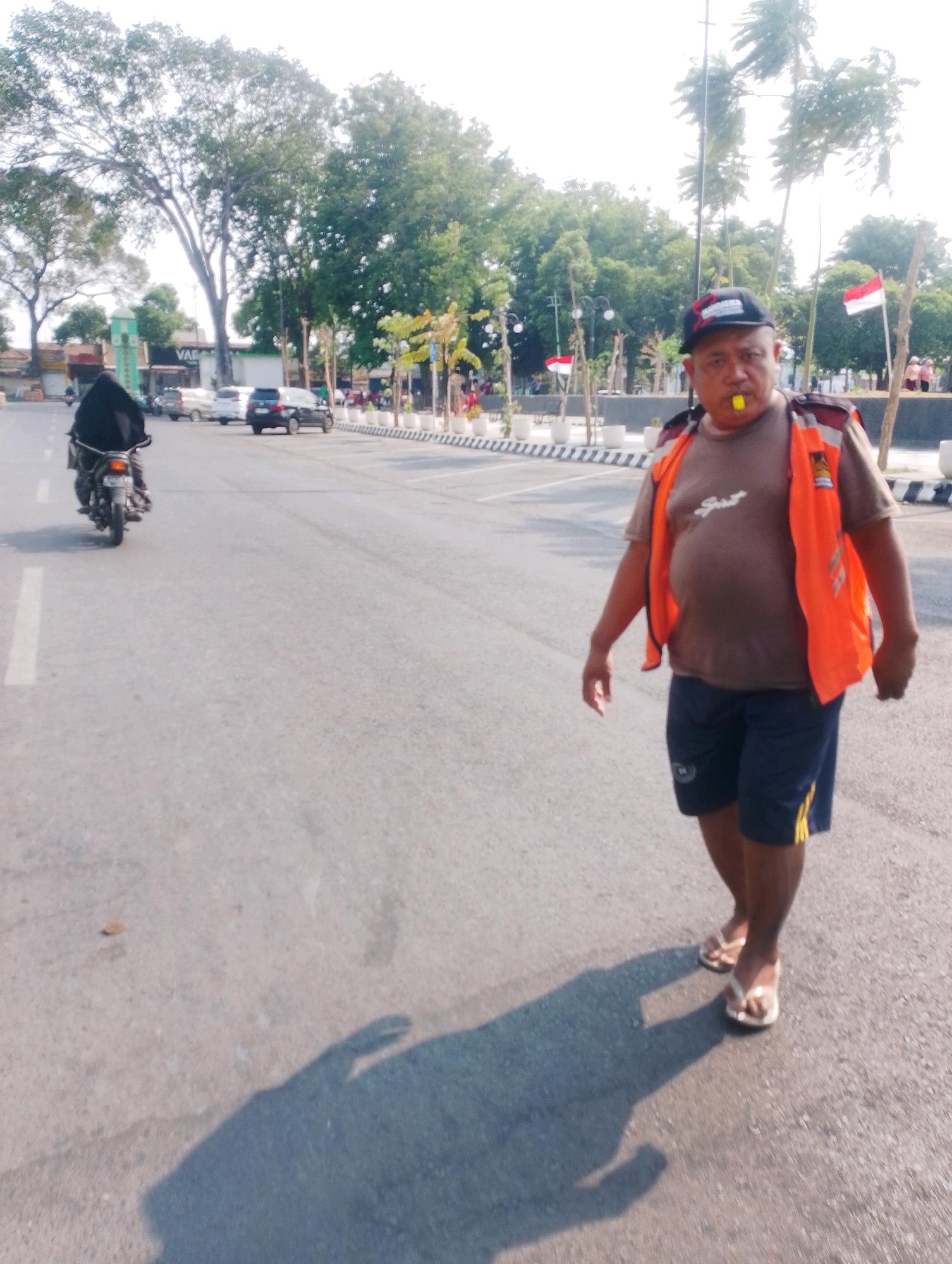 In Probolinggo, Indonesia, a man wearing an orange safety vest and a black baseball cap with a yellow whistle in his mouth walks on an asphalt road. He is wearing a brown t-shirt and dark blue shorts with yellow stripes. Behind him, a person on a motorcycle wearing a black head covering is visible. The road has white lane markings. In the background, there are trees, buildings, parked cars, and Indonesian flags. The sky is bright, suggesting it is daytime. The scene appears to be a street or road in a public area.
