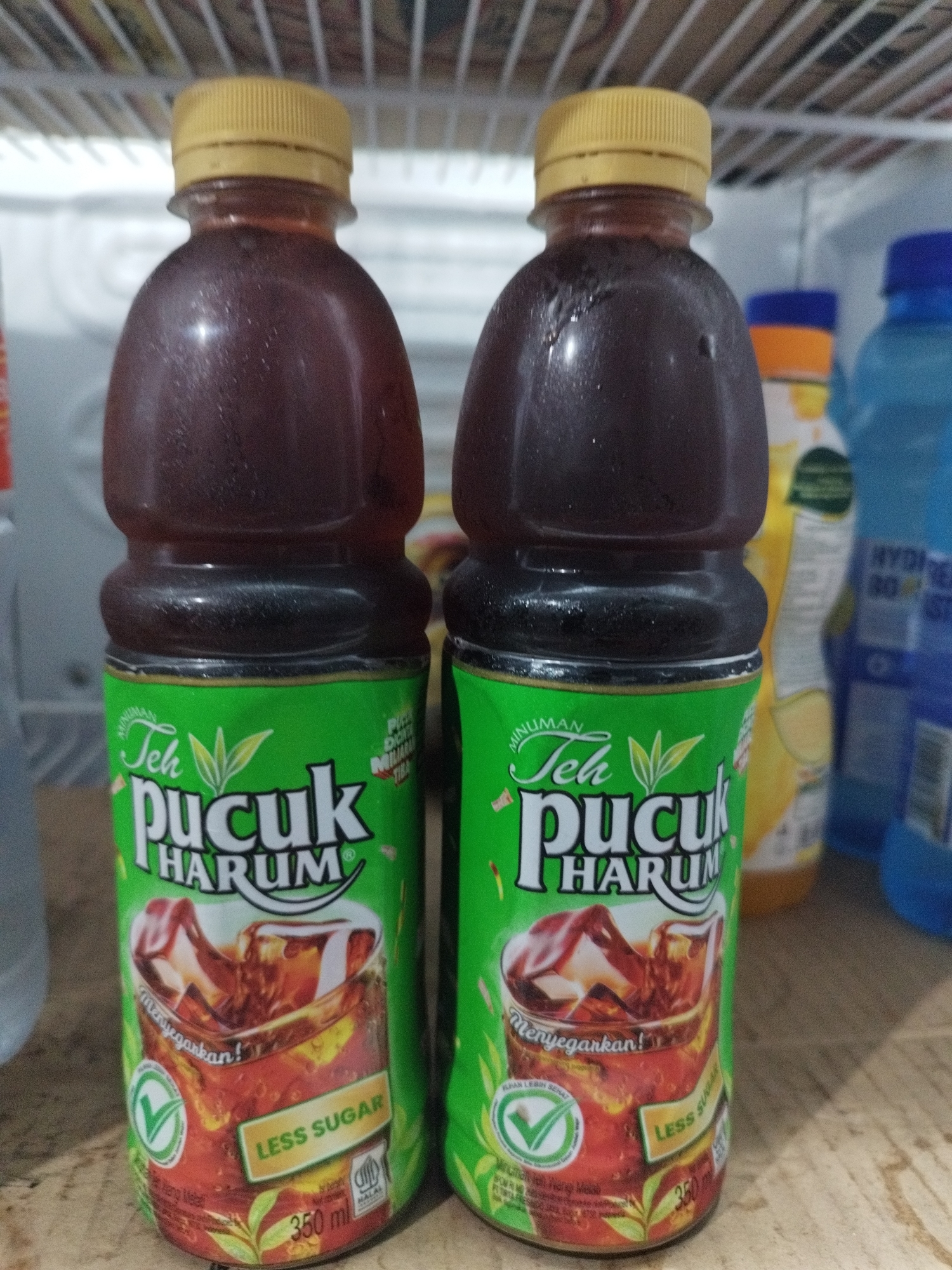 This is a close-up, eye-level, indoor, medium shot photograph of two bottles of Teh Pucuk Harum iced tea. The bottles are positioned in the center of the frame, side-by-side, with the left bottle slightly closer to the viewer. The liquid inside the bottles is a dark, reddish-brown color, and condensation is visible on the exterior of both bottles, indicating they are cold. Each bottle has a bright green label with white text. The main text on the label reads "Teh Pucuk Harum," accompanied by an illustration of a glass of iced tea. Below this, there is a smaller text that reads "LESS SUGAR" in a yellow rectangular box. A green circular logo with a checkmark is also present on the label, along with other smaller text and graphics. The bottle caps are yellow and ribbed.
The bottles are placed on a wooden surface, possibly a shelf or countertop, which has a visible grain pattern. In the background, other bottles of beverages are partially visible on shelving. To the right, a portion of a blue bottle and a yellow bottle are discernible. The shelving structure appears to be part of a refrigerator or display unit, with horizontal lines suggesting shelves. The overall lighting is bright, illuminating the subjects and their labels clearly. The image lacks any discernible signs of people, specific actions, or interactions. The environment appears to be a commercial setting, such as a minimarket or a refrigerator display area. While the image is a close-up of the product, it is not possible to definitively determine the exact location within the city of Karawang, Indonesia, as no specific landmarks or context are provided.
