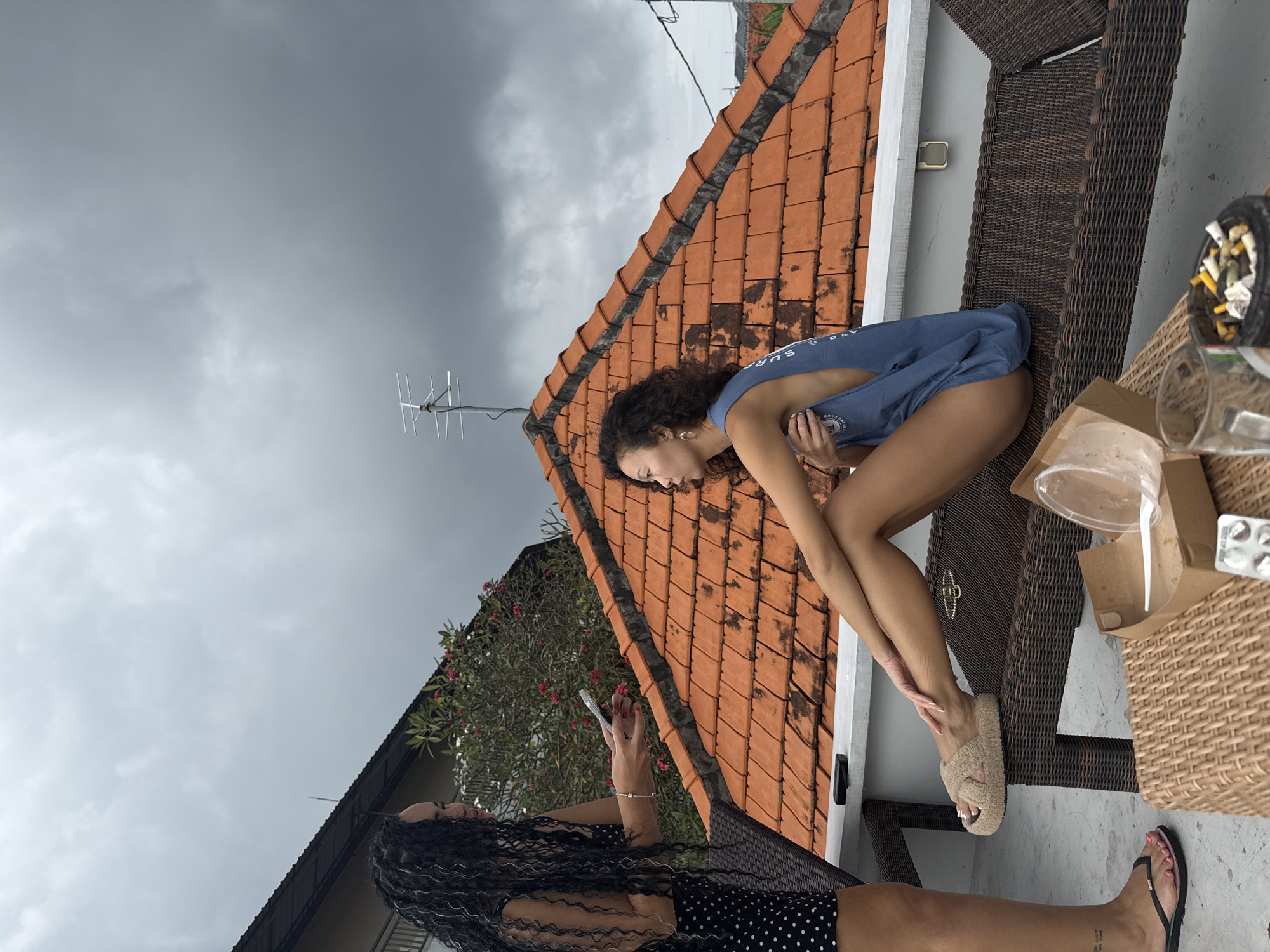 Two women are on a rooftop in North Kuta, Indonesia, under a cloudy sky. One woman, on the left, stands and wears a black dress with white polka dots and has long, dark, curly hair. She is holding a phone in her right hand, gesturing with it towards the other woman. The second woman is seated on a brown wicker lounge chair, facing left. She has curly, dark hair and is wearing a blue tank top. Her legs are crossed, and she is looking away from the standing woman. In the foreground, on a lower wicker table, there is a brown paper takeout container, a clear plastic cup, a glass mug with cigarette butts in it, and a blister pack of pills. The background features a terracotta tiled roof and a building.