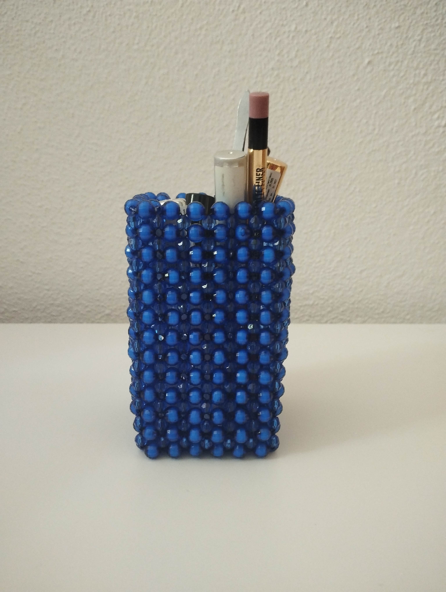 A decorative, rectangular container made of numerous small, shiny blue beads is centrally positioned on a flat, light-colored surface. The container, possibly a pen or makeup holder, exhibits a tightly woven pattern of the translucent, royal blue beads.
Inside the container, several items are stored upright. These include a light-colored pencil with a visible pinkish-brown tip, a metallic gold-colored tube, a silver or white cylindrical object, and a thin, white, pointed tool, possibly tweezers. The gold tube has black text on its side, clearly showing the word "EYELINER," indicating it is a cosmetic product.
The background consists of a light, off-white wall with a subtle textured finish. The lighting is bright and even, suggesting an indoor setting without direct sunlight. No specific activity is occurring; the image presents a still life of the beaded container and its contents. The overall impression is one of neat organization within a simple, clean environment.