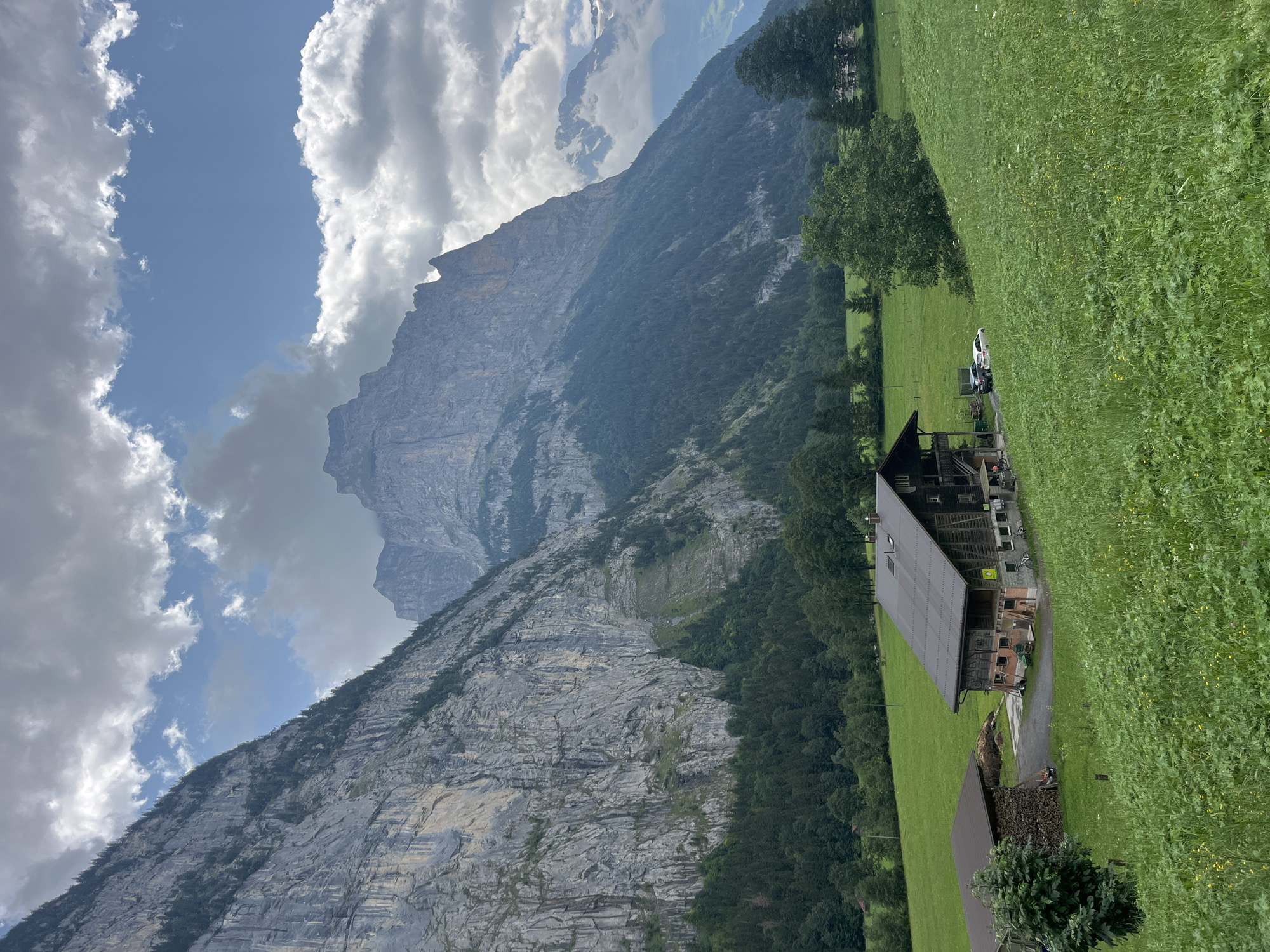 The image depicts a scenic view of the Lauterbrunnen Valley in Switzerland, characterized by towering, steep rock faces and lush green meadows. In the foreground, a traditional wooden barn with a dark metal roof sits in a verdant field dotted with wildflowers. Adjacent to the barn is a smaller structure covered by a dark roof, with a pile of firewood stacked next to it. Two vehicles, a dark-colored car and a white SUV, are parked near the barn. The middle ground is a sloping green pasture, and the background is dominated by immense, craggy mountains, their sheer rock walls textured and streaked with light and shadow. Patches of dark green conifer trees cling to the lower slopes of the mountains. The sky above is a mix of bright blue patches and heavy, grey clouds, suggesting a dynamic weather day. The overall scene conveys a sense of natural grandeur and rural tranquility.
