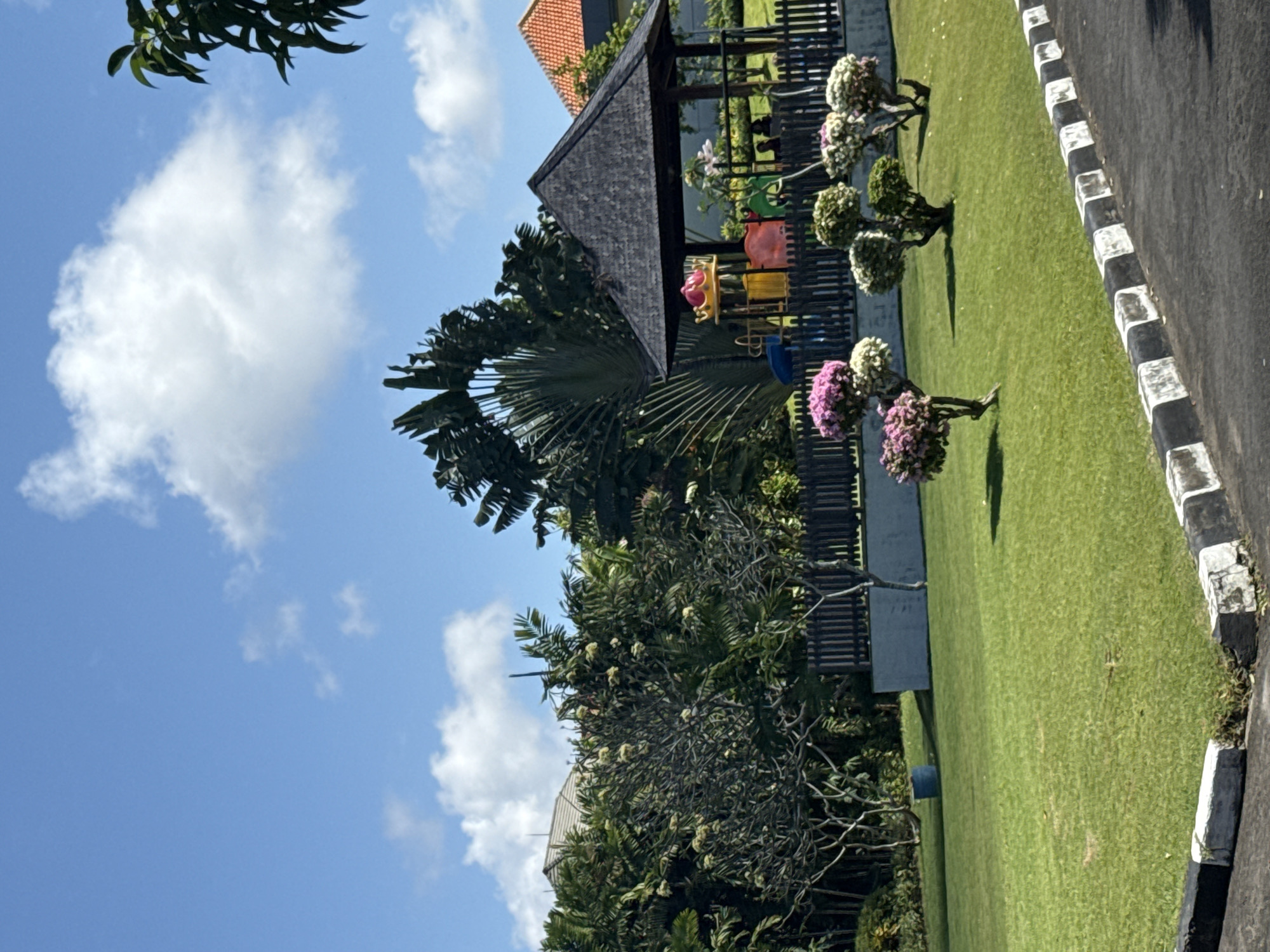 A bright, sunny day in Kuta Selatan, Indonesia, shows a well-maintained green lawn with several manicured bushes and trees. In the background, a dark-roofed gazebo stands next to a colorful playground structure, enclosed by a wooden fence. Beyond the fence, dense tropical foliage and a glimpse of a building with an orange-tiled roof are visible. A paved pathway with black and white striped borders runs along the bottom of the frame. The sky is a vibrant blue with scattered white clouds.