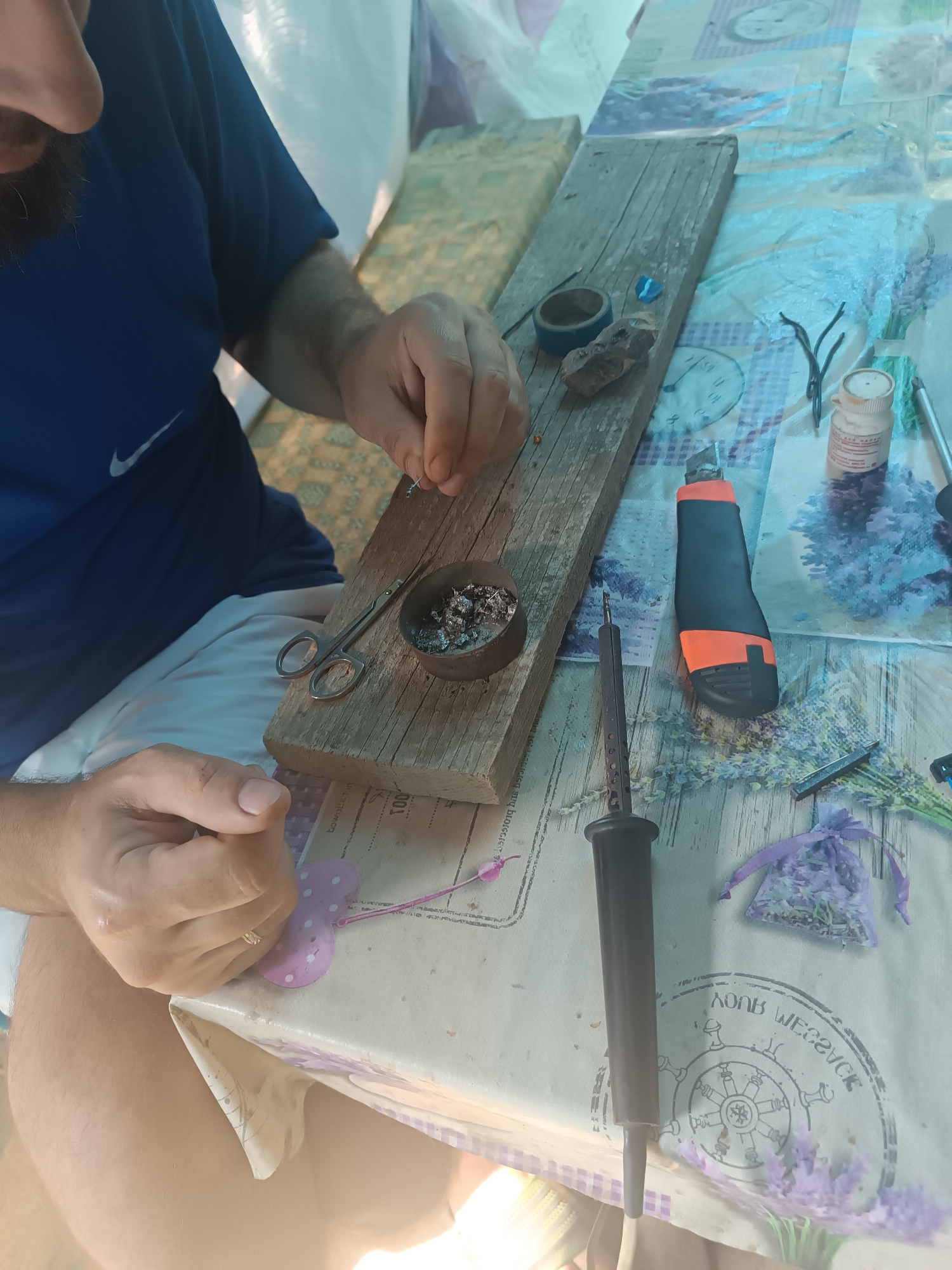In Vypasne, Ukraine, a person wearing a blue t-shirt with a white Nike logo is engaged in a craft activity at an outdoor table. The table is covered with a light-colored tablecloth featuring a lavender floral pattern and a circular nautical design. The individual is holding small metal pieces in their hands, seemingly working on a project.
In front of the person is a wooden plank on which various tools and materials are arranged. These include a pair of small scissors, a metal dish filled with shredded metal, a spool of dark tape, a rough stone, and a small blue object. To the right of the plank, a soldering iron with an orange and black handle and a black soldering tool are visible. Additionally, there is a small bottle with a red label, a bouquet of dried lavender, and a small purple sachet containing dried lavender. A retractable utility knife with an orange handle is also present on the table. The background suggests an outdoor setting, possibly a garden or backyard, with hints of white tenting or structures and dappled sunlight.