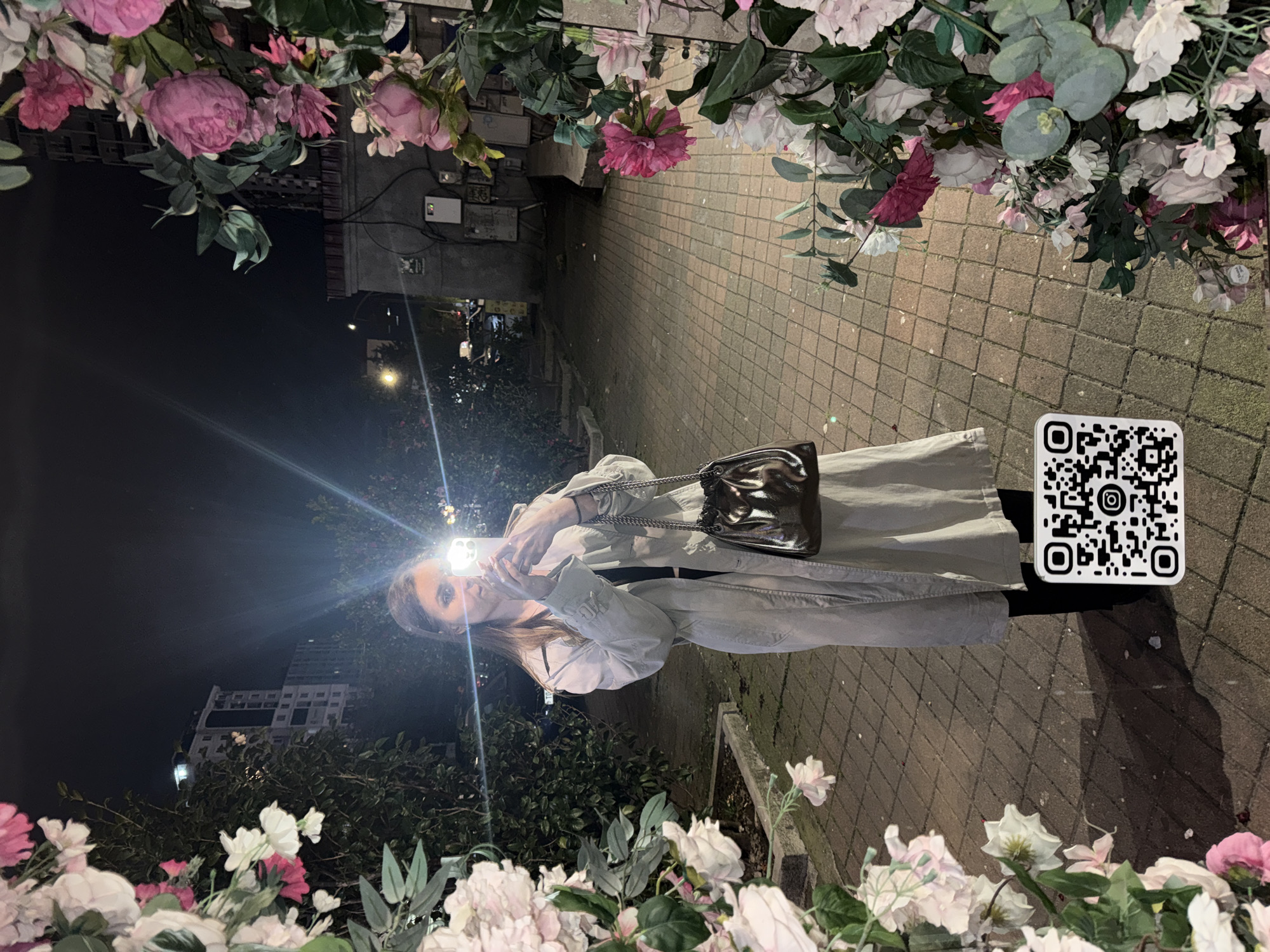 The image captures a young woman at night in what appears to be an urban setting in Batumi, Georgia. The woman, positioned centrally, is taking a selfie or video with her smartphone, which emits a bright light illuminating her face. She has brown hair and is wearing a long, light-colored coat or trench coat and a metallic or silver shoulder bag with a chain strap.
The scene is framed by decorative artificial flowers and foliage in shades of pink, white, and green, suggesting a thoughtfully adorned outdoor space. The ground beneath her is a paved surface with a brick or cobblestone pattern. In the background, under the dark night sky, outlines of buildings and distant artificial lights are visible, contributing to the evening ambiance. To the right of the woman, on the paved ground, there is a distinct black and white QR code printed on a white sticker. The overall mood is casual and modern, characteristic of a night out in a city.