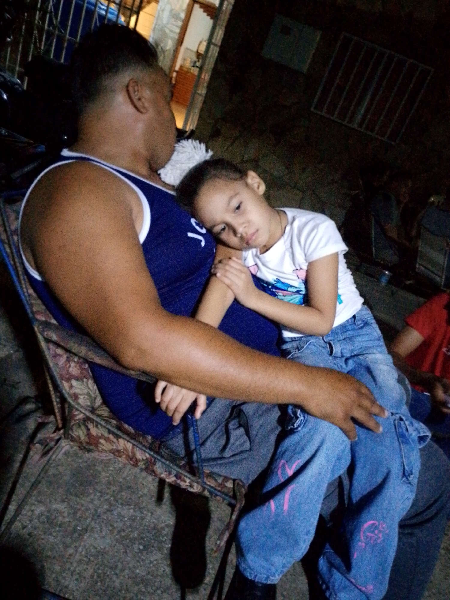 In this candid, dimly lit photograph, a man sits in a patterned outdoor chair, holding a young child on his lap. The man, with his arm around the child, wears a blue tank top. The child, leaning against the man, is dressed in a white t-shirt and blue jeans adorned with pink designs. The scene appears to be outdoors, possibly a patio or backyard, with concrete ground visible. In the background, a dark metal grate is mounted on a wall, and the faint glow of interior lights suggests it is evening. The overall mood is one of quiet repose, with the child seemingly resting or perhaps asleep. The location is identified as Juan Griego, Venezuela. There is no visible text other than the pink designs on the child's jeans.