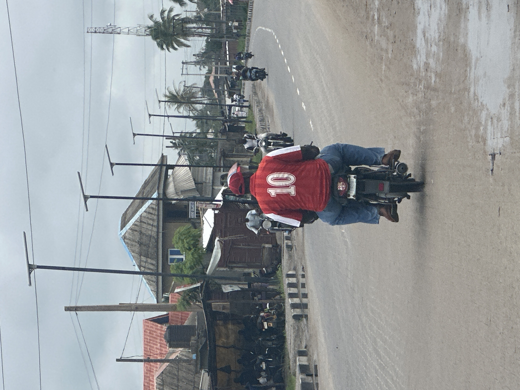 The image depicts a street scene in Badagry, Nigeria, under an overcast sky. A person wearing a red jersey with the number "10" in white, blue jeans, and brown shoes is riding a motorcycle from a rear perspective. They are positioned in the foreground, dominating the center of the frame. The motorcycle has a license plate with a crest.
The road appears to be paved, with a white dashed line indicating the lane. Several other motorcycles and riders are visible further down the road, some moving in the same direction and others in the opposite. To the left of the road, there are buildings with corrugated iron roofs, some appearing to be informal structures or shops, with a water tank visible. Beyond these structures, more distant buildings and vegetation, including palm trees, are present. Utility poles and electrical wires crisscross the sky. In the far background, a telecommunications tower can be seen. The overall environment suggests a bustling urban or semi-urban area.