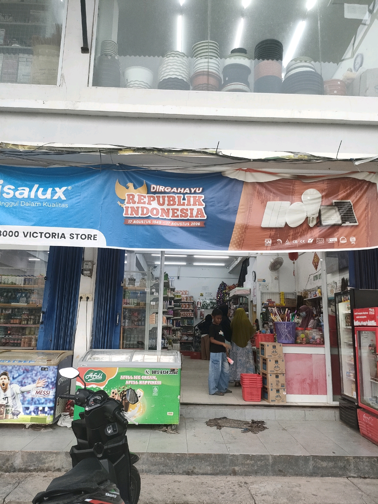 The image shows the exterior of a store in Batam, Indonesia. A blue and red banner hangs above the store's entrance, displaying the text "DIRGAHAYU REPUBLIK INDONESIA". Below the banner, the text "3000 VICTORIA STORE" is visible. The store appears to sell a variety of goods, including food, drinks, and household items, with shelves stocked with products visible through the open entrance. In front of the store, there are two refrigerators, one displaying an image of Lionel Messi and the other advertising "A FULL ICE CREAM". A black motorcycle is parked in the foreground. Several people are seen inside and near the entrance of the store, some appearing to be customers and staff. The environment suggests a commercial area with a mix of retail and possibly residential elements nearby, given the presence of overhead wires.