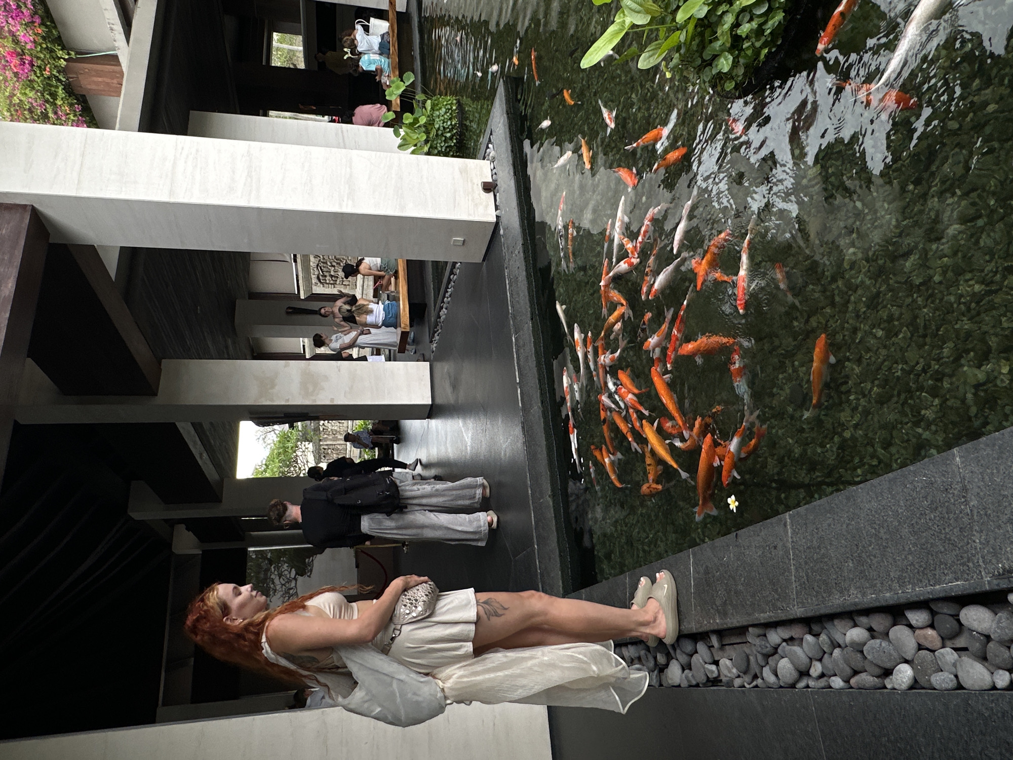 The image depicts a partially obscured view of a koi pond located in Nusa Dua, Indonesia. A woman with red hair, wearing a light-colored dress and slippers, stands beside the pond, holding a small silver purse. Other people are seen in the background walking through a building that is adjacent to the pond. The pond contains numerous orange and white koi fish. The edge of the pond is lined with smooth, gray pebbles. The environment includes architecture with dark and light pillars. The photograph is taken from a low angle.